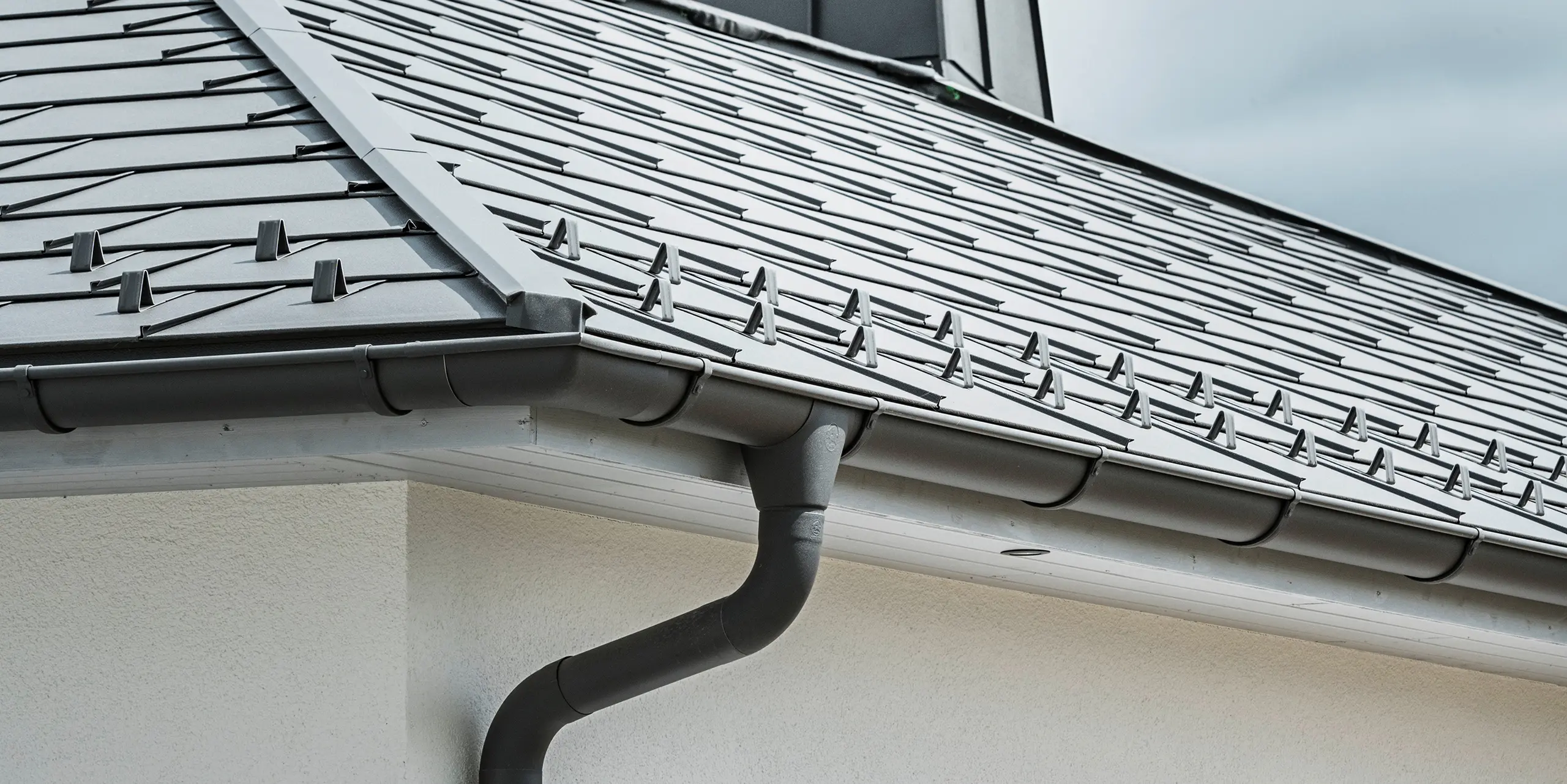 Close-up of the eaves of a detached house equipped with the efficient PREFA roof drainage system in P.10 dark grey. The image shows the detailed installation of the PREFA R.16 roof tile with the seamlessly integrated half-round gutter and downpipe underneath, which together provide a reliable and aesthetically pleasing solution for rainwater drainage. The robust aluminium products and careful installation underline the quality and durability of the PREFA complete system.