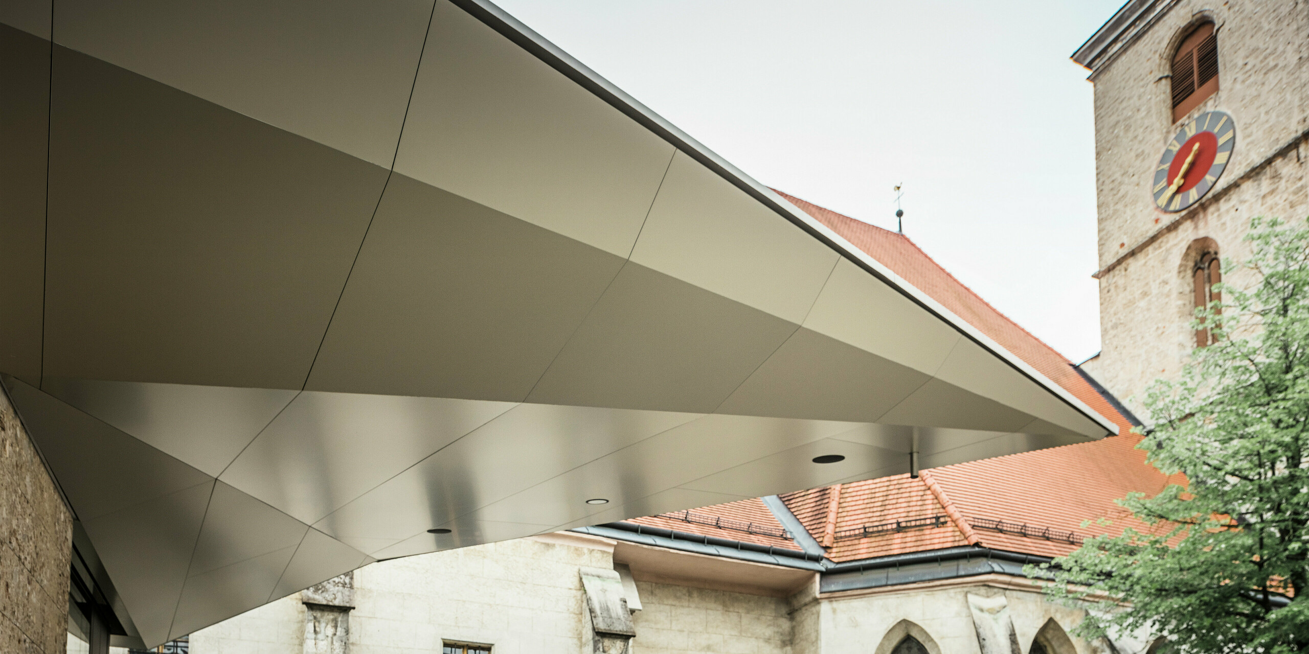 Dynamisch gespannte Dachfläche mit scharfkantiger Linienführung: PREFA Aluminium Verbundplatten ermöglichen diese präzise, fugenarme Gestaltung, die dem Eingangsbereich Leichtigkeit und Modernität verleiht - bei gleichzeitig hoher Witterungsbeständigkeit. Im Hintergrund ist die historische Kirche aus dem 14. Jahrhundert zu erkennen.