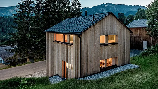 Aus erhöhter Perspektive zeigt sich Haus Immi eingebettet in die Berglandschaft von Ebnit. Warm beleuchtete Fenster setzen einen stimmungsvollen Kontrast zur naturbelassenen Holzfassade und zum schwarzen PREFA Solardach. Nahtlos integrierte Solardachplatten und Dachplatten R.16 in P.10 Schwarz verbinden effiziente Stromerzeugung, hohe Widerstandsfähigkeit und eine harmonische, moderne Dachgestaltung für alpine Architektur.