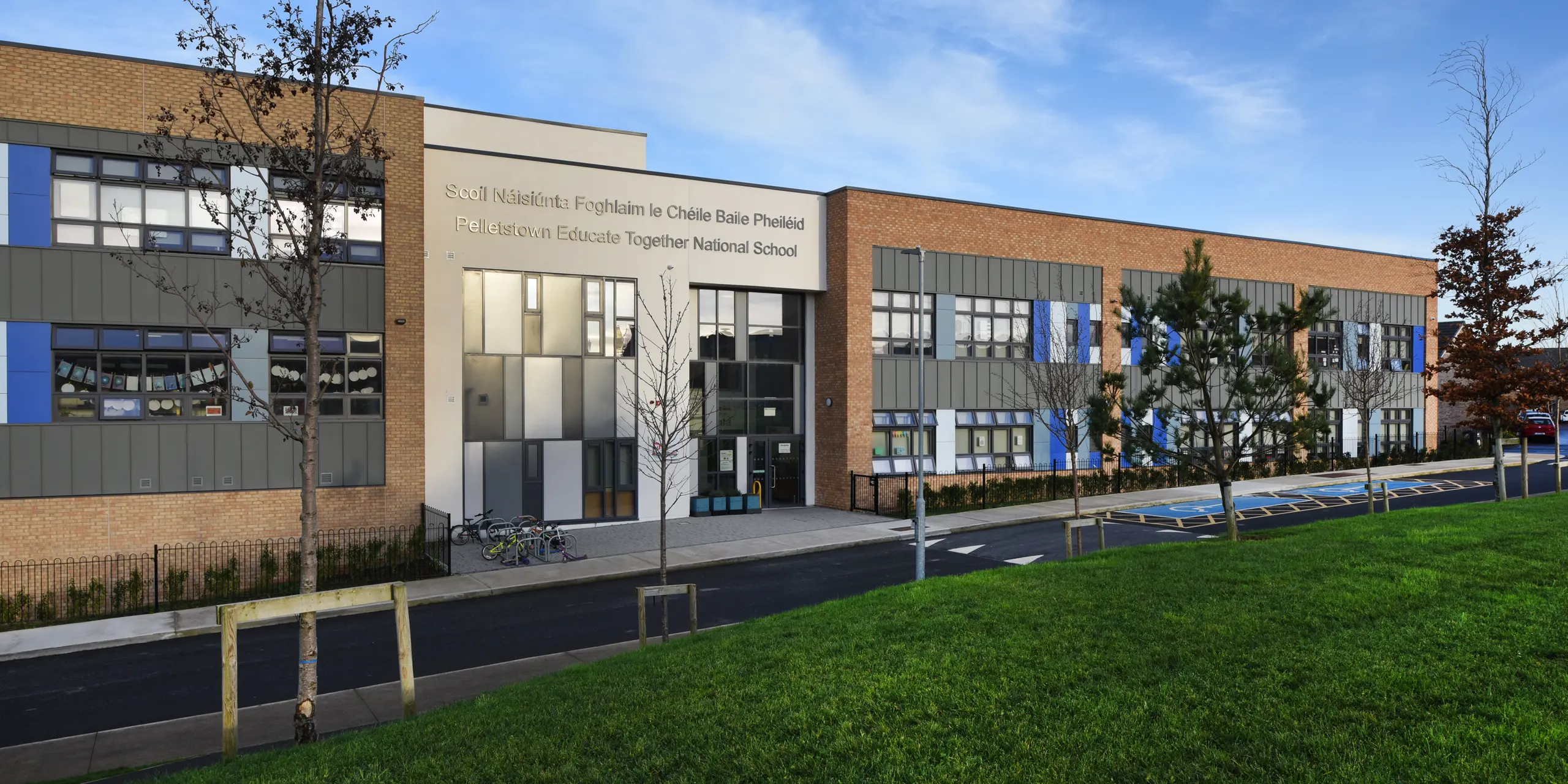 Front view of the Pelletstown Educate Together National School in Dublin with modern PREFALZ standing seam façade in P.10 light grey. The harmonious combination of aluminium, brick and plaster underlines the durable and elegant design of the school building. A well-tended green area with trees in the foreground emphasises the commitment to sustainability - as do the bicycles in front of the entrance.