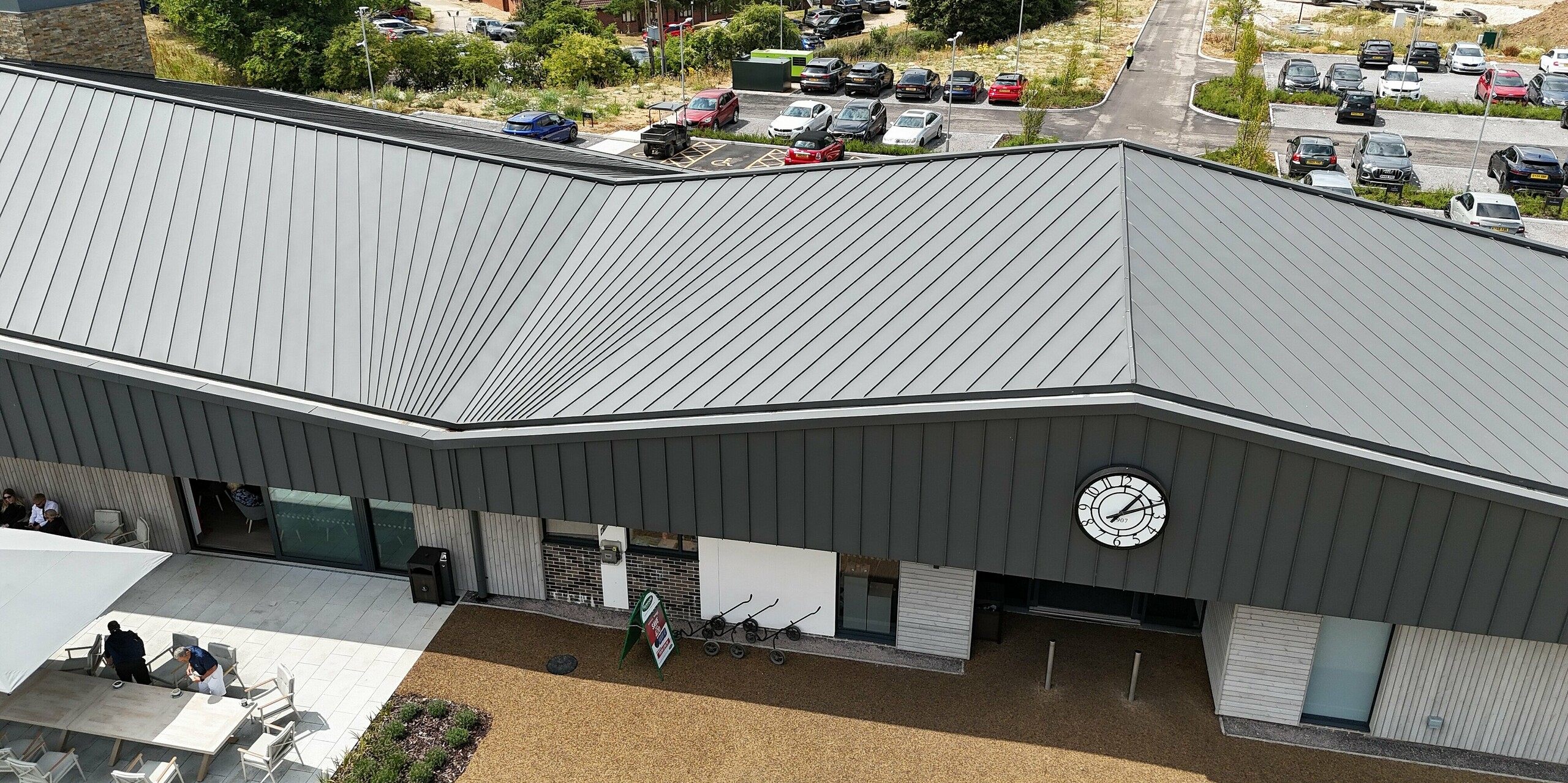 The impressive roof structure of the Basingstoke Golf Club in Hampshire, featuring PREFALZ standing seam panels in P.10 dark grey, immediately catches the eye. The curved shape of the roof and the precise arrangement of the standing seam panels create a striking, modern design that blends harmoniously into its surroundings. Wood panelling can be seen on the lower part of the façade, creating a natural contrast to the metallic surface. The outdoor seating area completes the picture of a modern, inviting clubhouse.