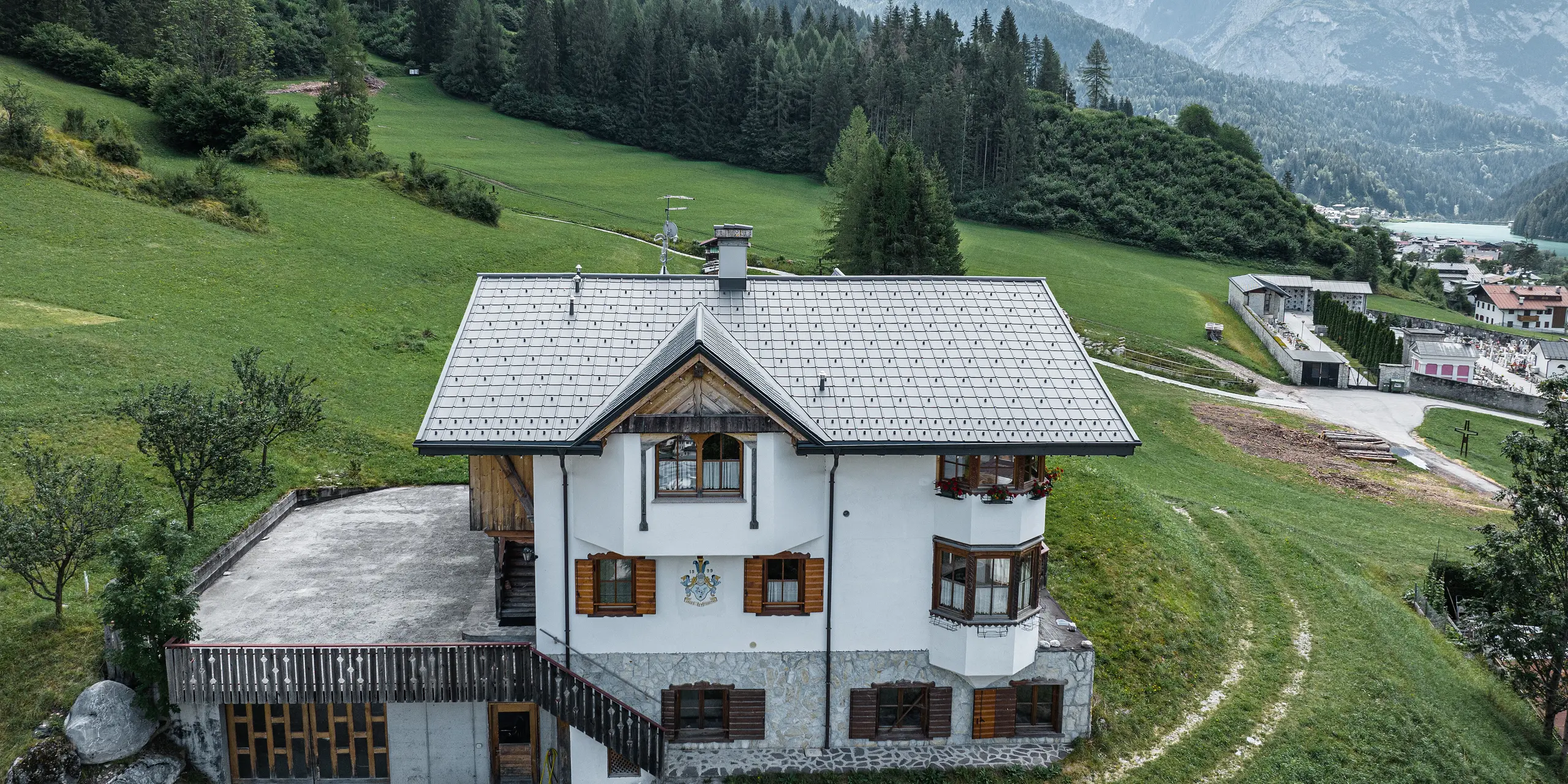 A bird's eye view of the entrance to the traditional mountain hut in Auronzo di Cadore after the roof renovation with PREFA tiles made of aluminium in P.10 light grey. The building, with its characteristic Alpine charm and modern sheet metal roof, blends harmoniously into the lush green landscape and the impressive mountain panorama in the background, demonstrating a successful combination of functionality and regional architecture.