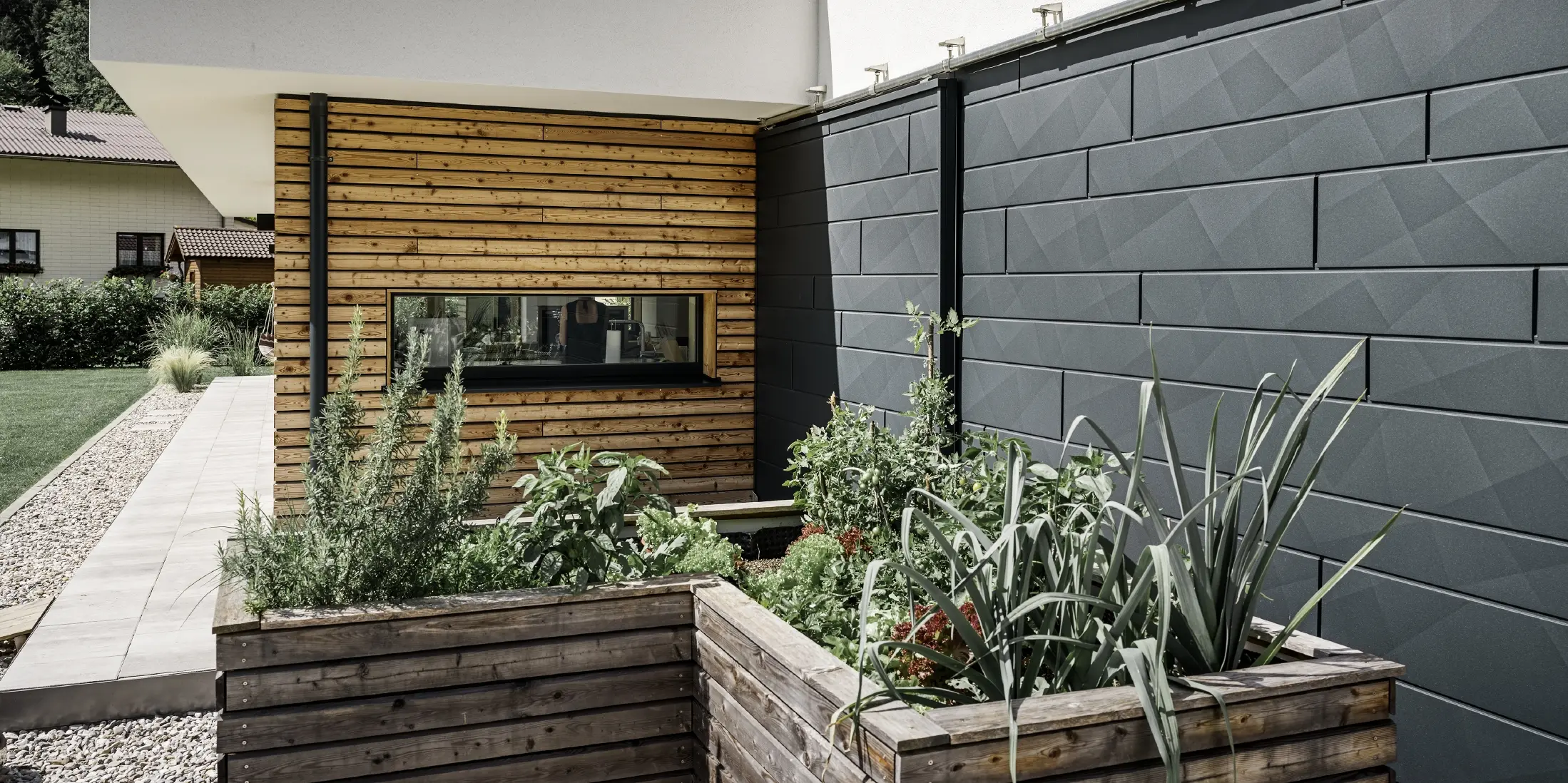 This image shows a close-up of the detached house, focusing on the façade cladding made from PREFA Siding.X in P.10 anthracite. The dark façade panels, with their distinctive crinkled finish, provide an appealing visual contrast to the vibrant, timber-clad areas and the surrounding green spaces. Raised beds, planted with various plants and shrubs, are positioned in front of the aluminium façade, emphasising the house’s natural and sustainable character. The combination of modern materials and integration into the garden area conveys an image of contemporary architecture that blends harmoniously into its surroundings.