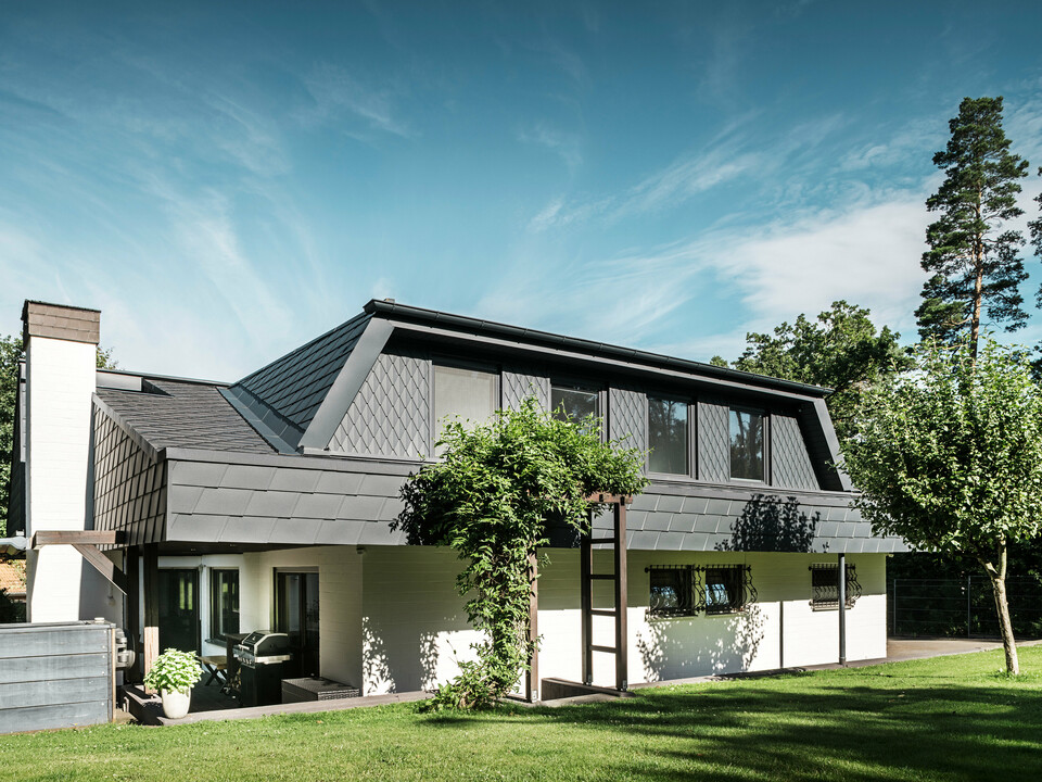 Seitliche Ansicht eines Einfamilienhauses in Mechernich mit PREFA Dachschindeln DS.19 und kleinen Dachrauten in P.10 Anthrazit. Die Trapezgauben sind mit Dachrauten aus robustem Blech verkleidet, während das Dach mit Schindeln gedeckt ist. Eine grüne Pergola und Bäume im Gartenbereich ergänzen die moderne und wetterbeständige Architektur.