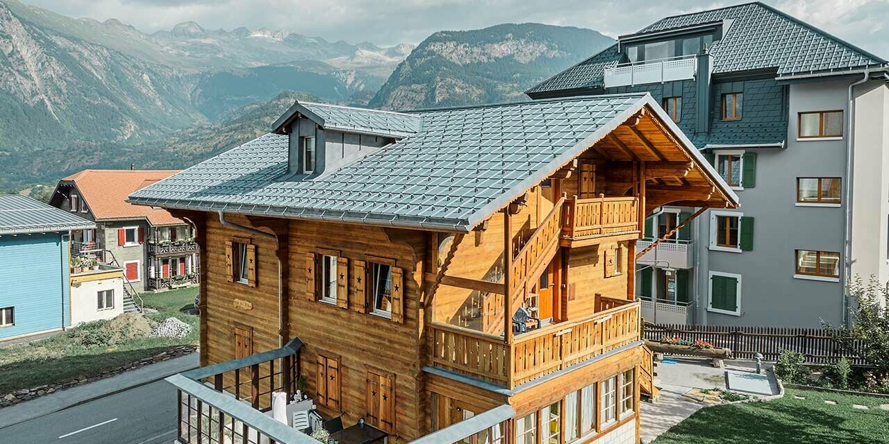 Traditional Swiss wooden chalet with dormer and gabled roof. The roof is clad in the classic PREFA roof tiles in stone grey.