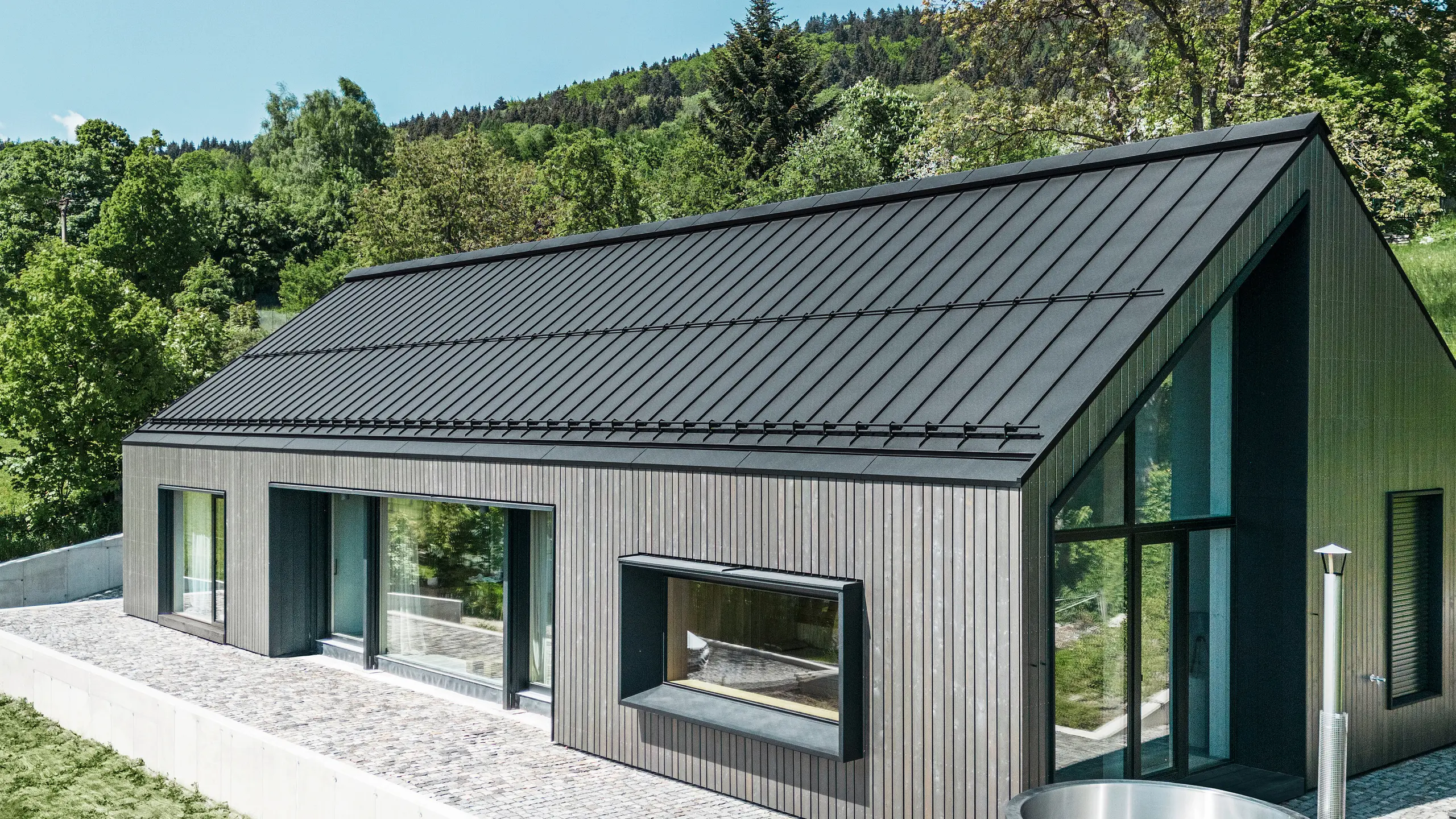 A modern wooden house in Kuncice (CZ) was equipped with large windows. The dark wooden house has a PREFALZ (standing seam) roof system in the colour P.10 black. In the rear outdoor area, there is a hot tub with wooden panelling.