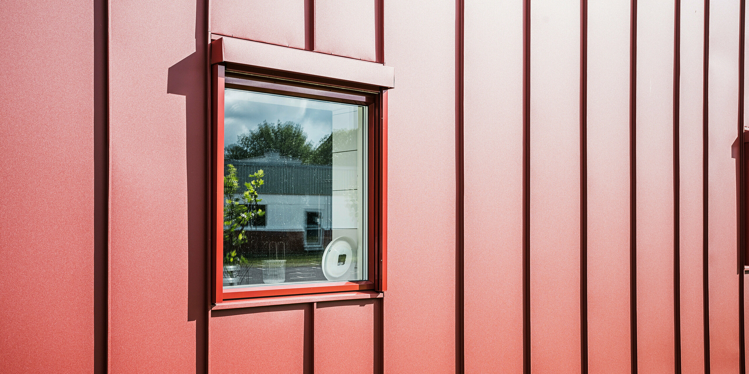 Nahaufnahme eines Fensters der Schulkantine Gröna Hjärtat in Broby, Schweden, zeigt die detailreiche PREFALZ Fassade in Oxydrot. Die präzise Verarbeitung der Stehfalzdeckung aus Aluminium unterstreicht die hohe Qualität und Langlebigkeit der Gebäudehülle. Das Fenster ist harmonisch in die Fassade integriert und reflektiert die Umgebung, was die moderne und klare Architektur des Baukörpers betont. Das ansprechende Design macht das Bauwerk zu einem Beispiel moderner und nachhaltiger Bauweise.