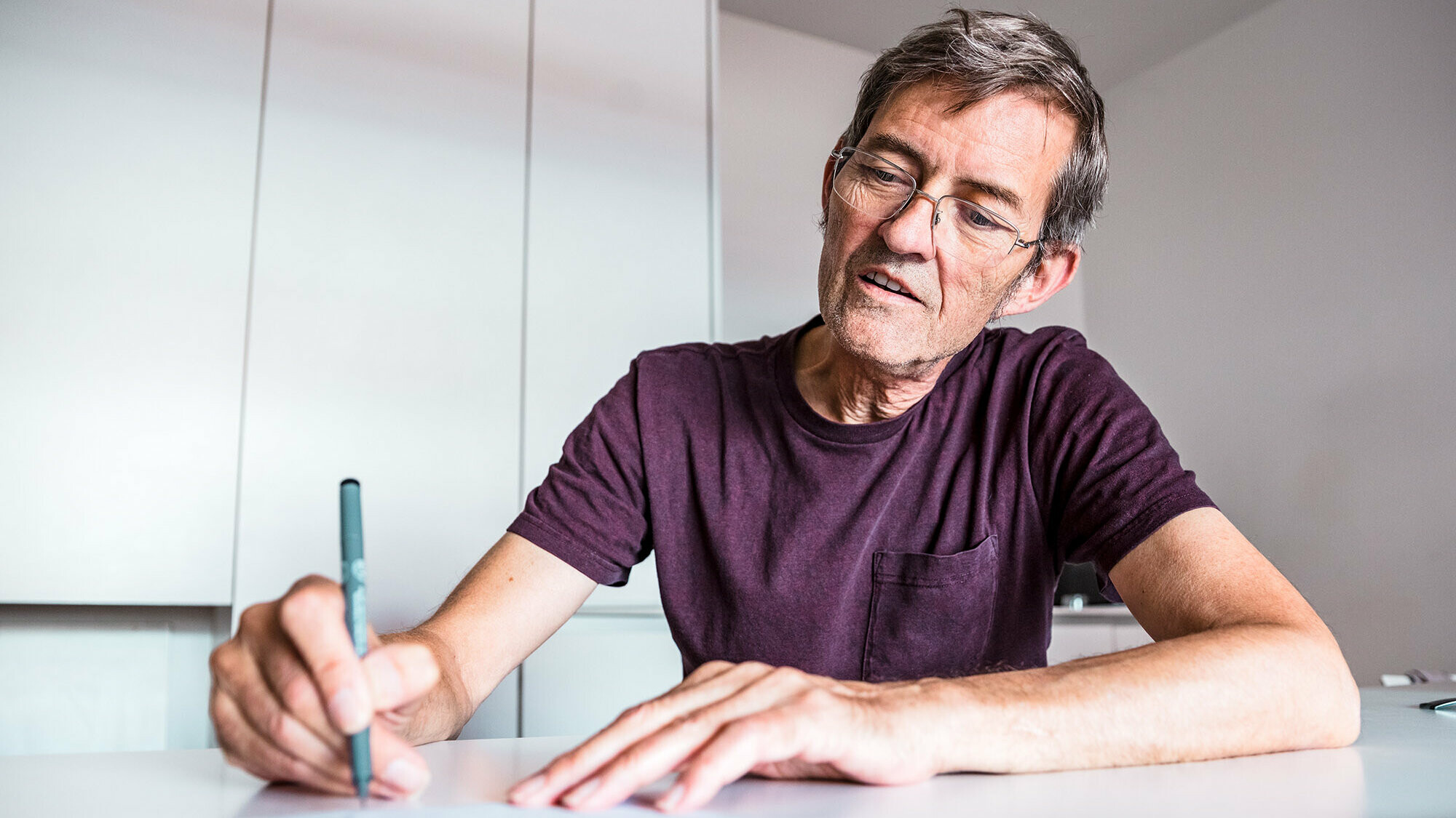 Architect Dominique Cornaert sitting at a white table and drawing; behind him, white walls as well as a white cupboard can be seen.