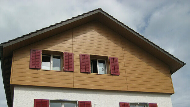 Gable cladding of a traditional house with a gable roof. The gable is clad with PREFA natural oak sidings, installed horizontally. The windows have red shutters.