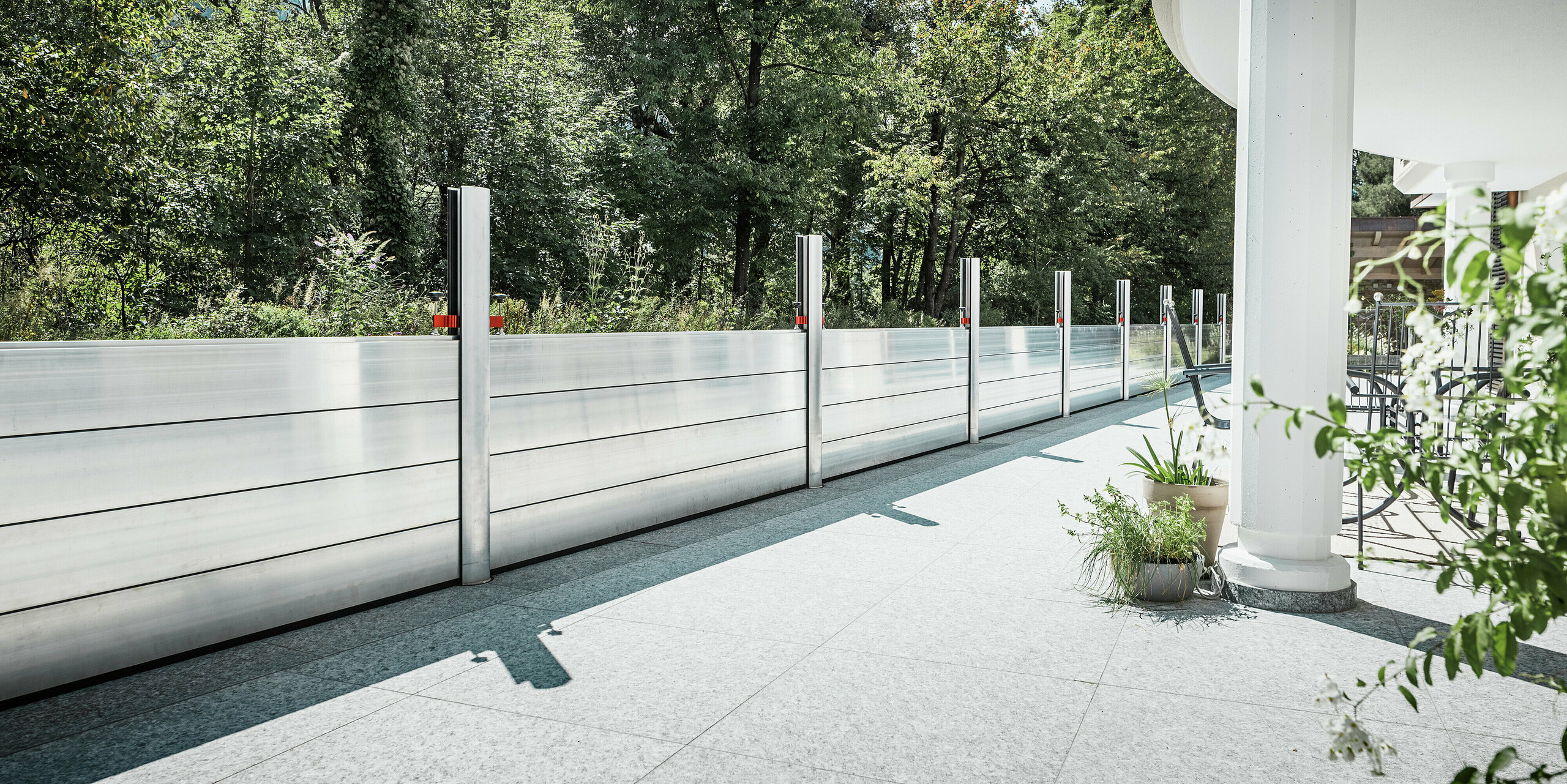 Blick von einer elegant geschützten Terrasse mit installiertem PREFA Hochwasserschutzsystem. Die Dammbalken aus Aluminium verlaufen geradlinig entlang der Grundstücksgrenze zum angrenzenden Wald und bieten einen effektiven Hochwasserschutz. Die glänzenden Aluminiumelemente fügen sich harmonisch in das moderne Design des Wohnhauses ein. Im Vordergrund: weiße Säulen, Pflanzkübel und Gartenmöbel, die die wohnliche Atmosphäre unterstreichen.