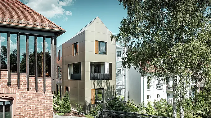 The image shows the modern town hall in Alzenau, whose façade is made of bronze-coloured aluminium composite panels from PREFA. The pointed roof and clearly structured windows with wooden blinds give the building an elegant and contemporary aesthetic. In the foreground is a green area with a paved path and trees, which harmoniously complements the urban design of the building.