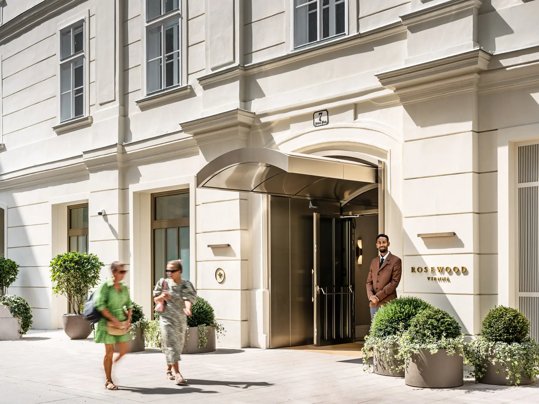 Entrance to the Rosewood Hotel with a porter, passers-by and larger potted plants in front of it.