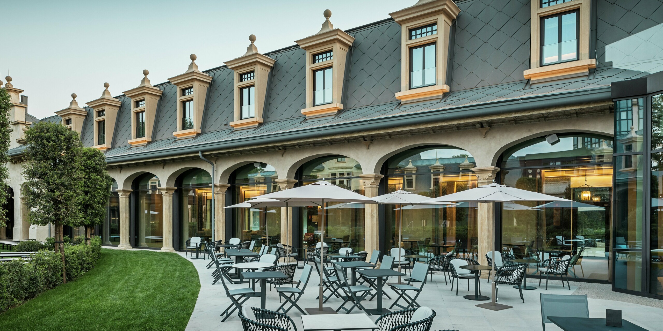 Die stilvoll gestaltete Terrasse des Thermalparks „De Montel – Terme Milano“ lädt mit modernen Sitzgruppen und Sonnenschirmen zum Verweilen ein. Im Hintergrund trifft denkmalgeschützte Architektur auf moderne Funktionalität: Das Aluminiumdach mit PREFA Dachrauten 44 × 44 in P.10 Hellgrau fügt sich harmonisch in die fein restaurierte Jugendstilfassade ein. Großflächige Glasfronten schaffen Transparenz und verbinden Innen- und Außenraum auf elegante Weise.