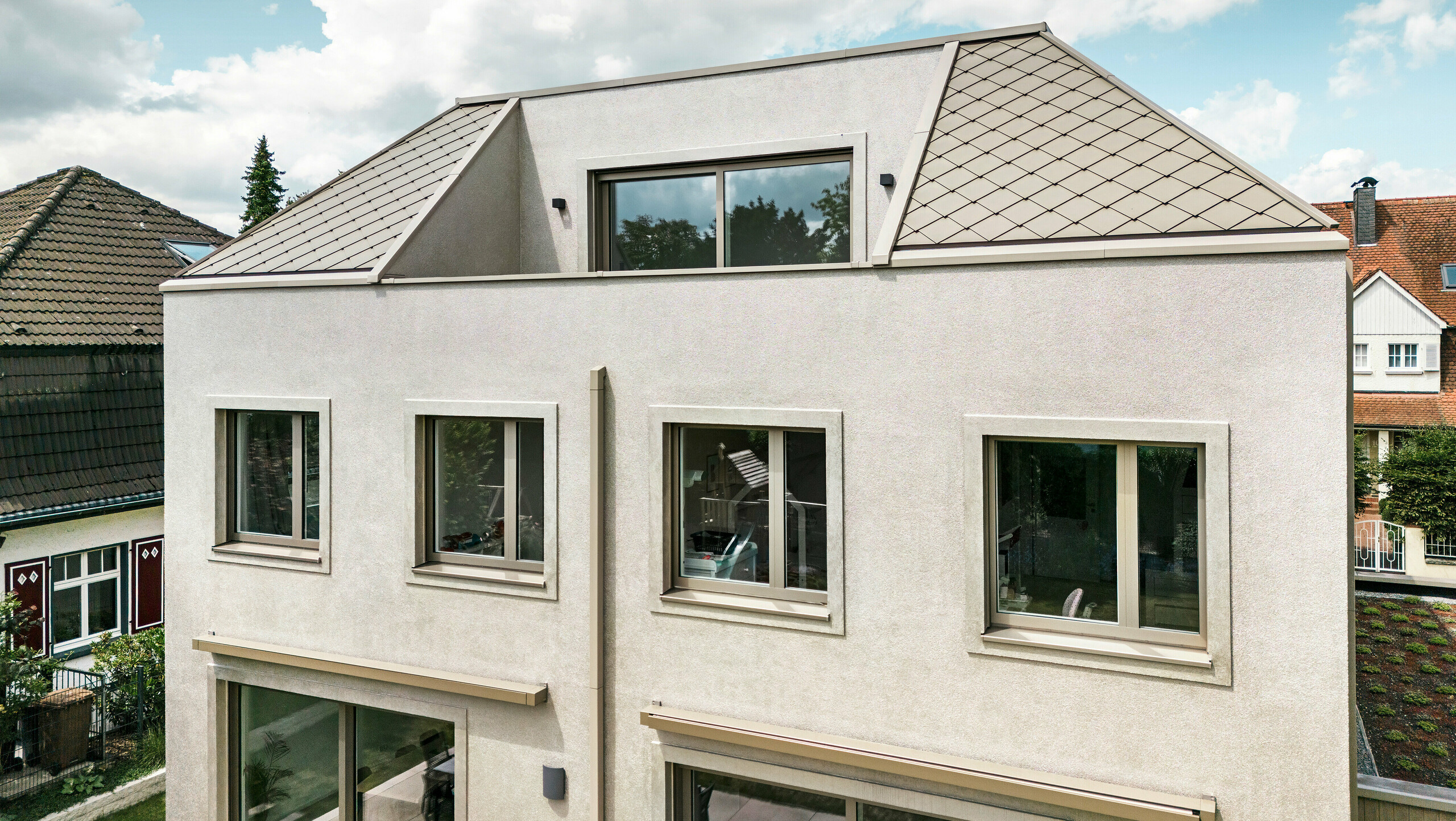 View of the upper floor of a modern detached house with a hipped roof and bronze-coloured aluminium roof cladding from PREFA. The roof is covered with elegant aluminium rhomboid tiles, which underline the contemporary and stylish design of the house. The upper floor has large windows that let in plenty of natural light. The façade of the house has a simple and modern design, giving the building an elegant and harmonious look. Other houses in the neighbourhood can be seen in the background, emphasising the harmonious overall appearance of the residential area.