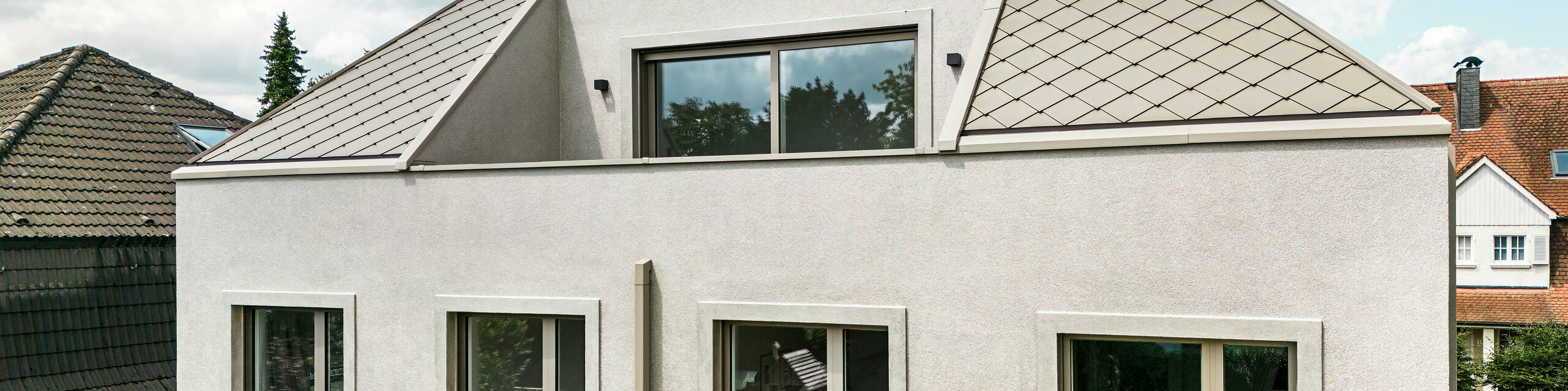 View of the upper floor of a modern detached house with a hipped roof and bronze-coloured aluminium roof cladding from PREFA. The roof is covered with elegant aluminium rhomboid tiles, which underline the contemporary and stylish design of the house. The upper floor has large windows that let in plenty of natural light. The façade of the house has a simple and modern design, giving the building an elegant and harmonious look. Other houses in the neighbourhood can be seen in the background, emphasising the harmonious overall appearance of the residential area.