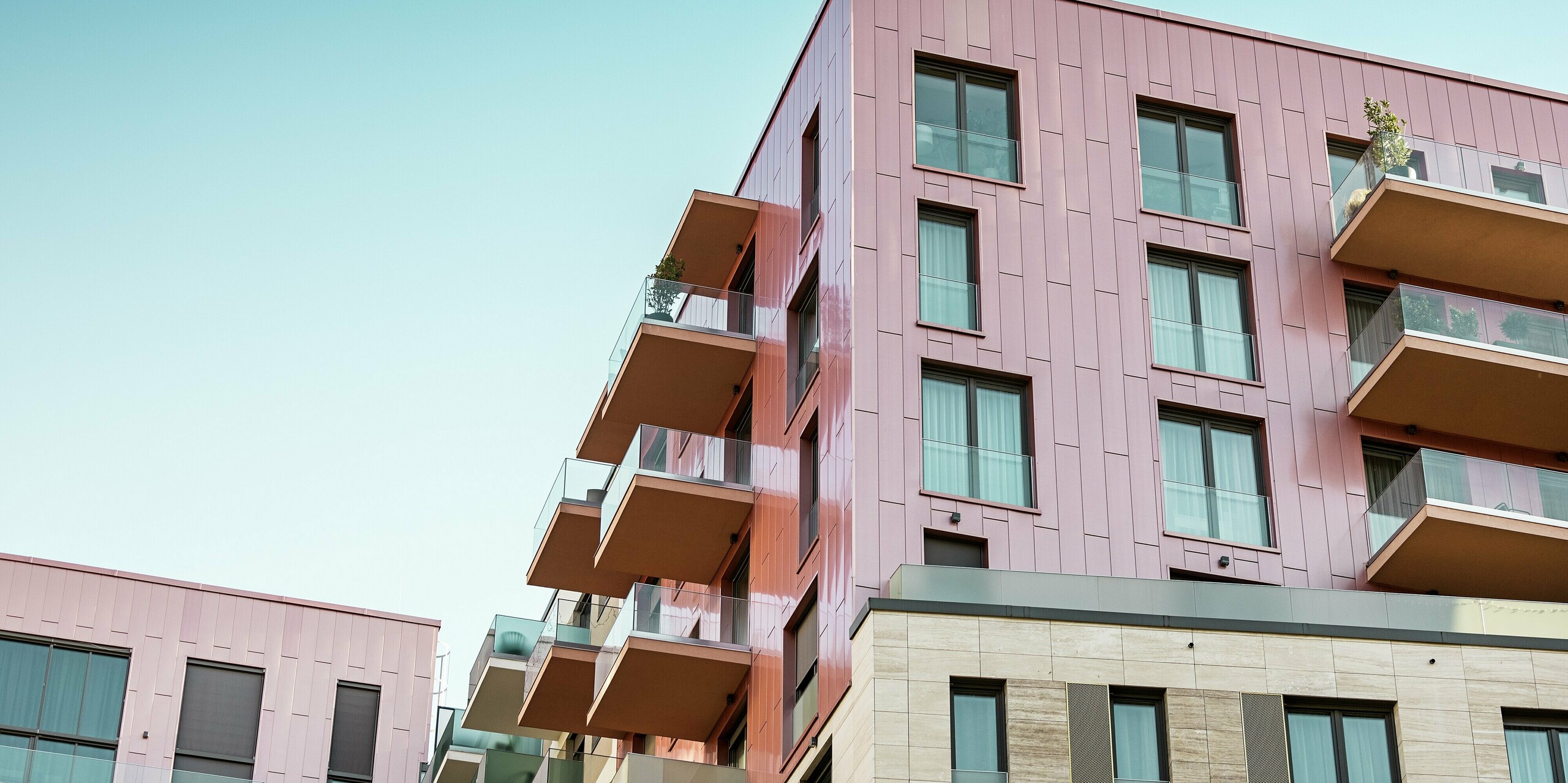 Modernes Wohngebäude im Budapester Corvin-Viertel mit auffälliger PREFA Siding Fassade in vertikaler Verlegung und der Sonderfarbe Lachsmetallic, gebürstet. Die präzise verlegten Aluminiumfassadenpaneele betonen die klare Linienführung des Baukörpers und erzeugen ein hochwertiges, urbanes Erscheinungsbild. Die langlebige PREFA Fassade steht für moderne Architektur, Witterungsbeständigkeit und Wartungsfreiheit im Stadtraum.