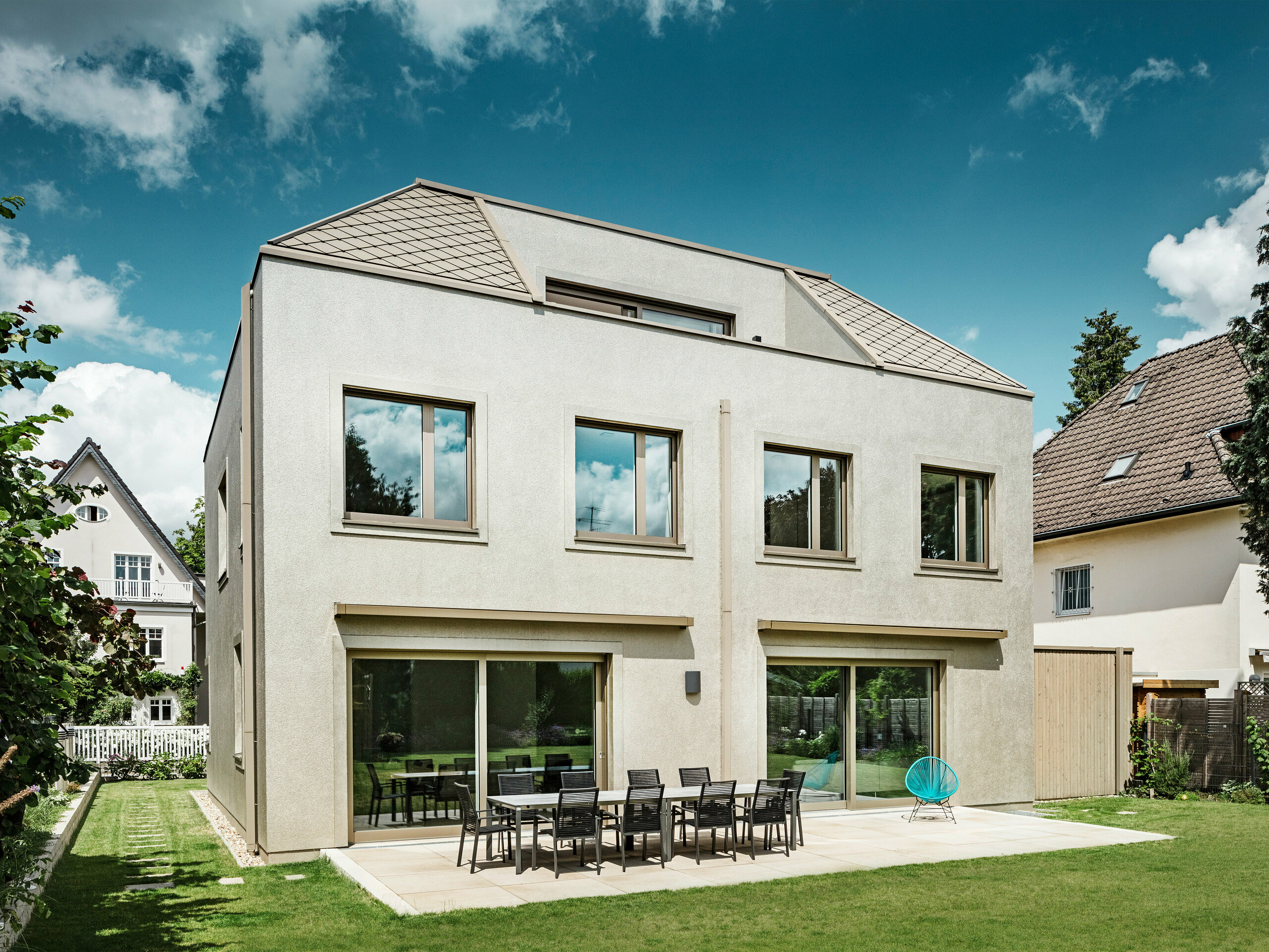 A modern detached house with a hipped roof and bronze-coloured aluminium roof and façade cladding from PREFA. The house is located in a quiet residential neighbourhood, surrounded by well-tended gardens and other detached houses. Large windows can be seen in the background, flooding the living area with plenty of natural light. A spacious terrace with garden furniture invites you to relax and linger. The design of the house blends harmoniously into its surroundings, while the modern choice of PREFA materials gives it a contemporary and elegant look.