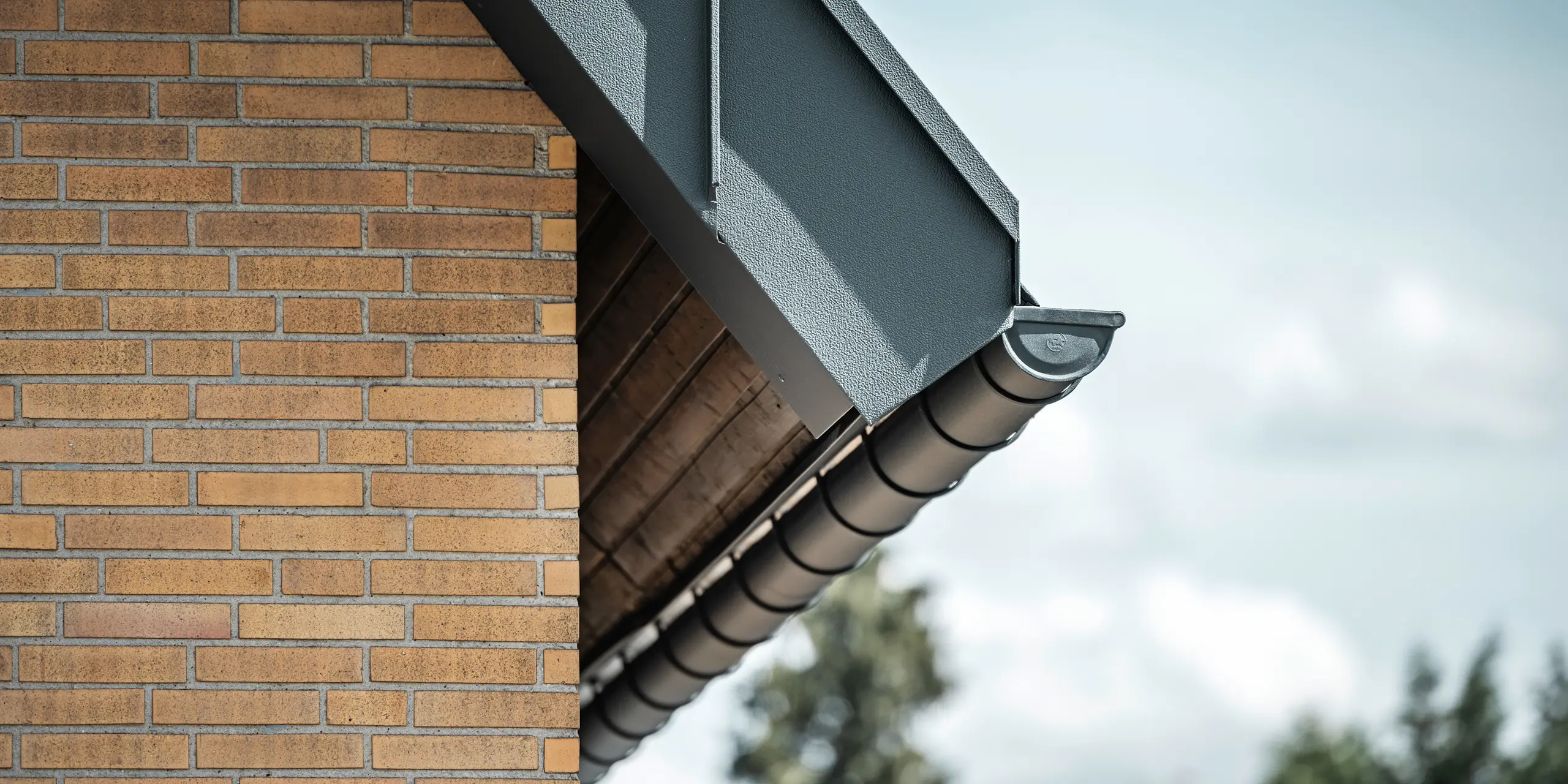 Detailed view of an anthracite-coloured roof drainage system from PREFA. The half-round gutter stands out clearly against the warm tones of the brick wall and emphasises the precise workmanship and quality of the materials used.