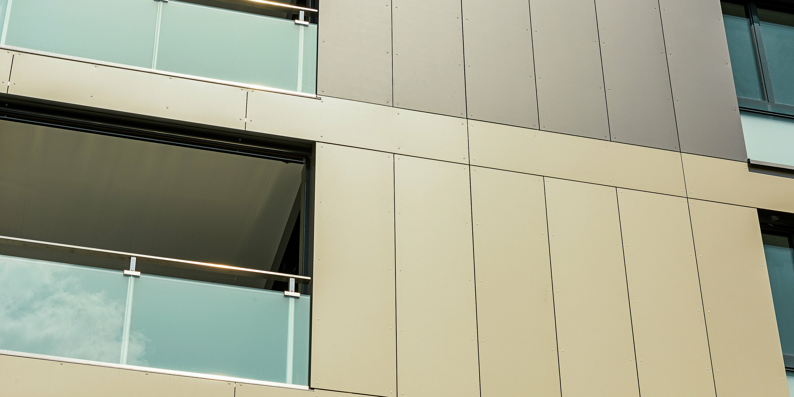 Nahaufnahme der Aluminiumfassade an der kubischen Wohnanlage in Murten (CH). Die Aluminium Verbundplatten von PREFA garantieren eine hohe Widerstandsfähigkeit und Langlebigkeit. Die harmonische Farbgestaltung in Bronze und Schwarzgrau fügt sich perfekt in die Umgebung ein und schützt das Gebäude effektiv vor äußeren Einflüssen wie Wind und Wetter. An den Bildrändern sind Balkone mit Glasverkleidungen zu sehen.
