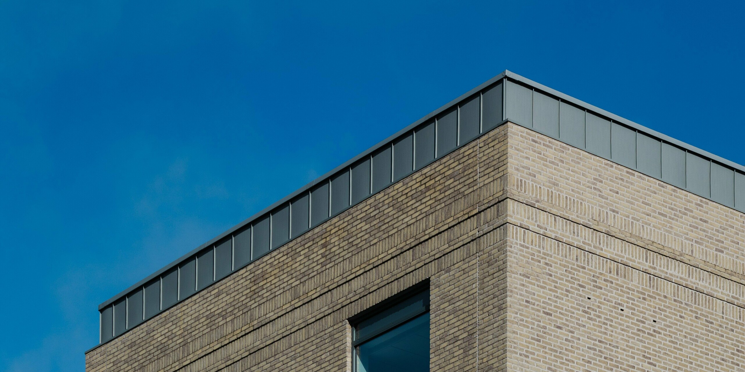 The Northern Regional College in Ballymena stands out as a contemporary educational building with sharp geometry and refined material contrasts. Light brickwork is complemented by vertically installed PREFALZ standing seam aluminium panels in patina grey, forming a striking architectural highlight at roof level. PREFA aluminium offers durability, weather resistance and a low-maintenance surface. The subtle grey tone contrasts beautifully with glass elements and blue sky, emphasizing the building’s precision, longevity and modern architectural elegance.