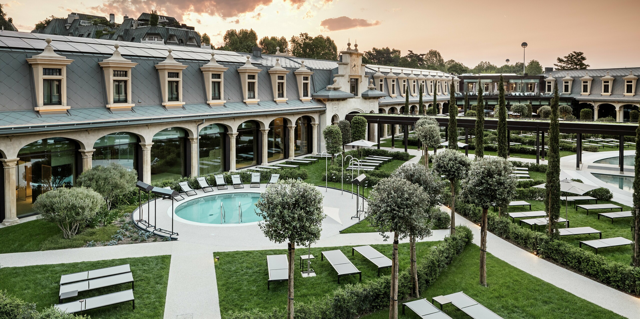 Abendstimmung im Thermalpark „De Montel – Terme Milano“ mit eindrucksvollem Blick auf die denkmalgeschützte Fassadenarchitektur im Jugendstil. Die Dacheindeckung mit PREFA Dachrauten 44 × 44 in P.10 Hellgrau verbindet Nachhaltigkeit mit Ästhetik. Im Vordergrund laden Liegewiesen, gepflegte Wege, Olivenbäume und ein rundes Thermalbecken zum Verweilen ein.