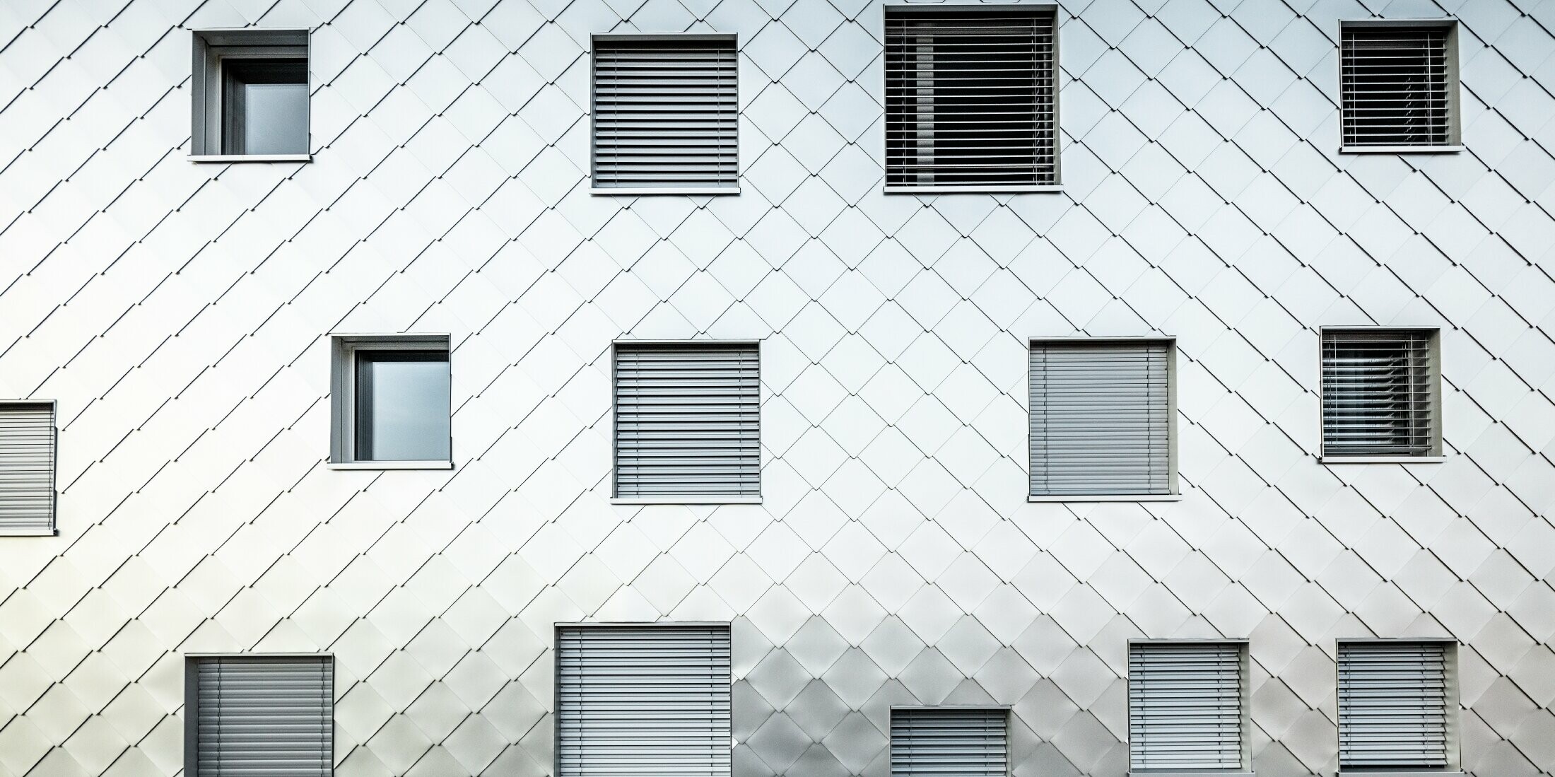 Das Bild zeigt eine Nahaufnahme der Fassade des Solitaire-Hochhauses in Horw, Luzern, das von der Architektin Tilla Theus entworfen wurde. Die Fassade ist mit eloxierten Aluminiumrauten verkleidet, die ein diagonales Muster bilden. Die Fenster sind unregelmäßig angeordnet und tragen zur einzigartigen Optik des Gebäudes bei. Einige Fenster sind mit Jalousien ausgestattet. Die Oberfläche der Aluminiumrauten reflektiert die Umgebung und verleiht der Fassade eine dynamische und glänzende Erscheinung.