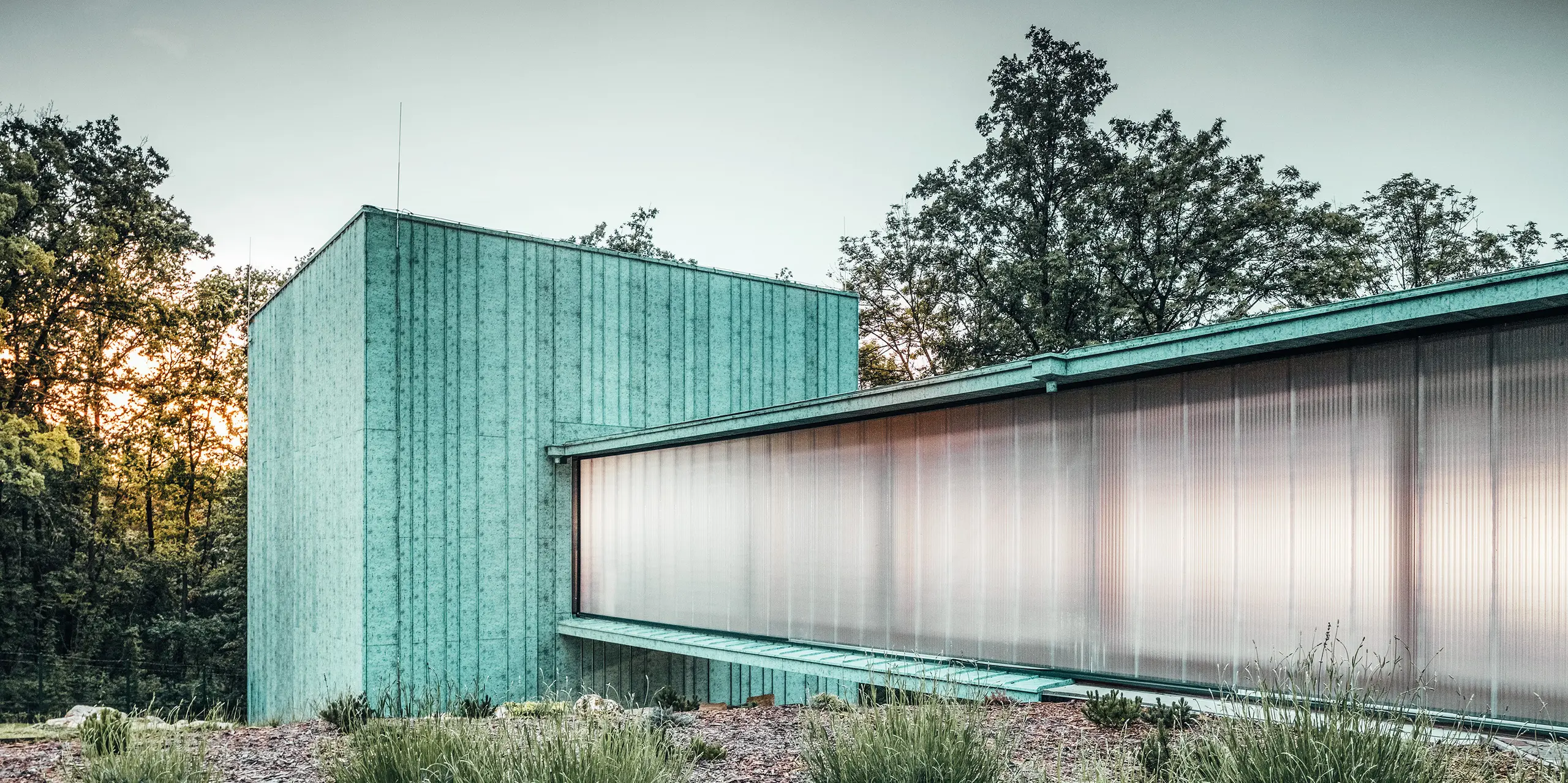 Das Zero Magnetic Field Laboratory in Fertőboz (HU) ist umhüllt von PREEFALZ in der Farbe P.10 Patinagrün. Das würfelförmige Laborgebäude durch einen Tunnel mit dem Nebengebäude verbunden. Die Seitenwände des Durchgangs verleihen dem Bauwerk eine Transparenz durch ihre Milchglasfolierung. Im Hintergrund ist die natürliche Landschaft im Licht des Sonnenuntergangs zu sehen. 