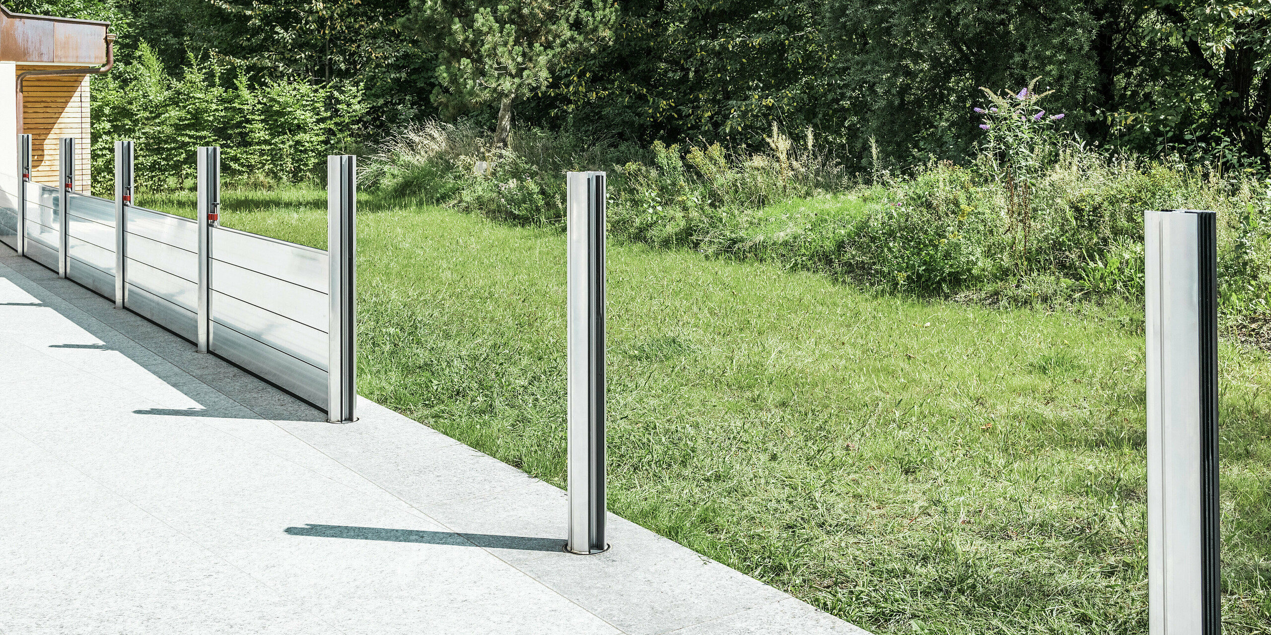 Detailansicht des mobilen PREFA Hochwasserschutzsystems beim Aufbau auf einer Terrasse in Tirol. Zu sehen sind bereits montierte Bodenhülsen sowie eingesetzte Aluminiumstützen, die das Dammbalkensystem aufnehmen. Im Hintergrund erkennt man die angrenzende Wiese und das offene Gelände - ein typisches Einsatzgebiet für den PREFA Landschaftsschutz. Die hochwertige Aluminiumkonstruktion bietet zuverlässigen Schutz vor Hochwasser bei eleganter Optik. In den beiden vorderen Stützen sind noch keine Dammbalken eingesetzt, da sich das System zum Zeitpunkt des Fotos in der Aufbauphase befindet.