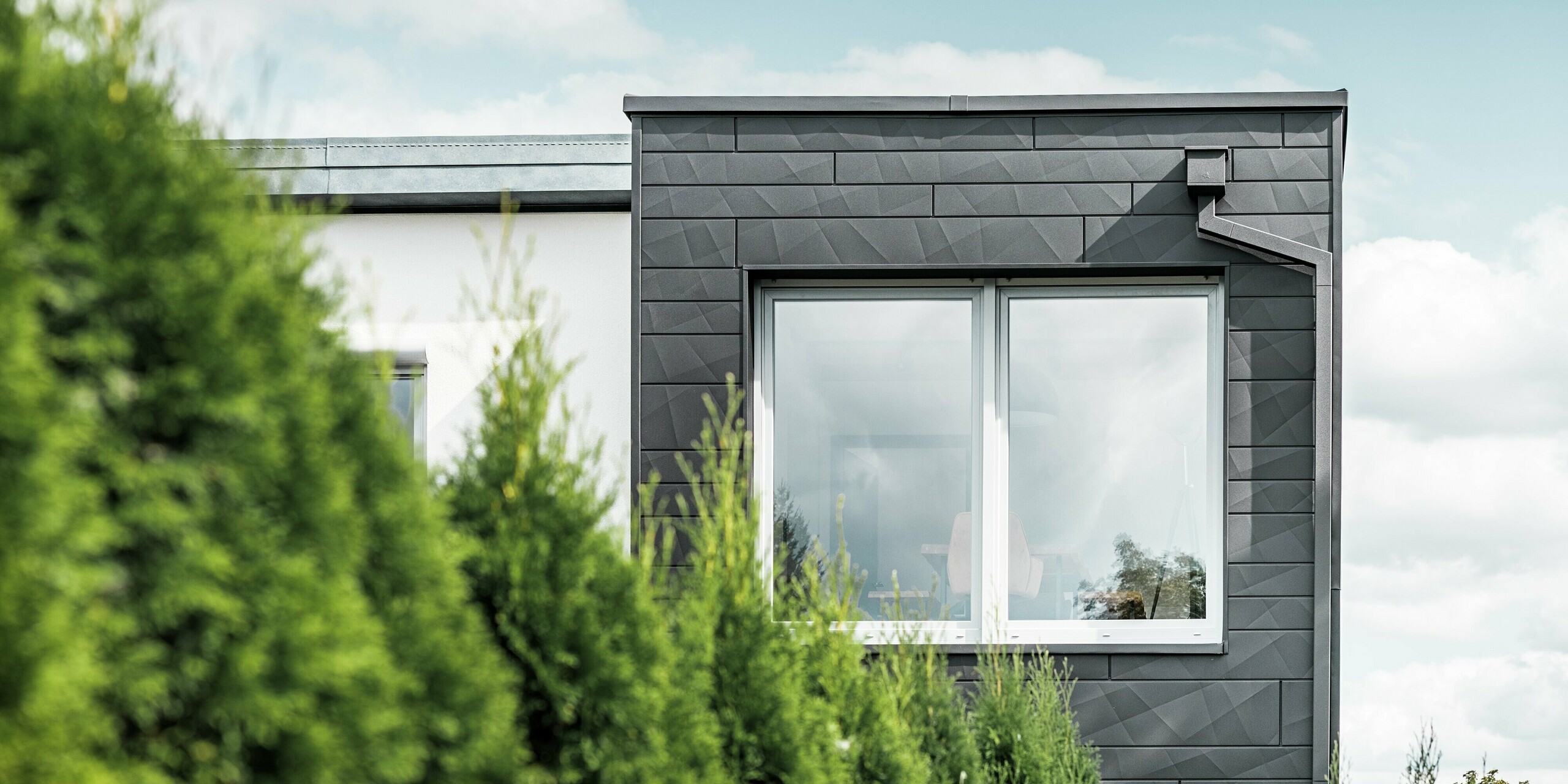 The clearly structured PREFA siding.X façade panels in colour P.10 black adorn the exterior wall of a single-family home in Teuschnitz, giving it a unique appearance. The PREFA roof drainage system, consisting of the square downpipe and the leader head, blends seamlessly into the design. The green thuja trees in the foreground contrast with the dark sheet metal facade.