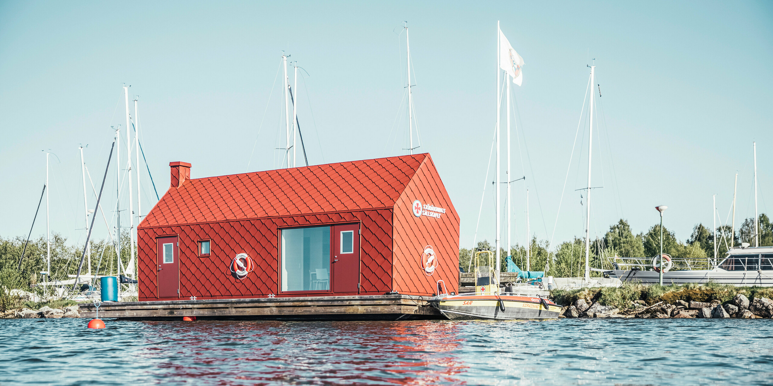 Das schwimmende Stationshaus der Seenotretter am Ufer des Hjälmaren Sees, umgeben von ruhigem Wasser und Segelbootmasten, erstrahlt in der Sonne Schwedens. Die lebendige, oxydrote Aluminiumverkleidung aus PREFA Dach- und Wandrauten 29×29 bietet nicht nur optische Wärme, sondern auch strukturelle Integrität, um den harten klimatischen Bedingungen zu trotzen. Ausgestattet mit Rettungsringen und positioniert als ein Wächter der Sicherheit, symbolisiert dieses Gebäude die Zuverlässigkeit und Bereitschaft der Seenotretter, denen es als Heimat dient.