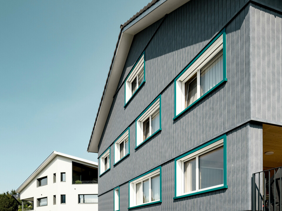 Ansicht zweier benachbarter Mehrfamilienhäuser in Weissbad, Schweiz, wobei das vordere Gebäude mit seiner PREFA Siding Fassade in P.10 Steingrau einen starken visuellen Eindruck vermittelt. Die vertikal montierten Sidings schaffen eine elegante und schützende Hülle, während die türkisen Fensterrahmen farbliche Akzente setzen. Das Bild fängt das Zusammenspiel der modernen Materialien mit der traditionellen Architektur ein und demonstriert die vielseitige Einsetzbarkeit von PREFA Aluminiumprodukten.