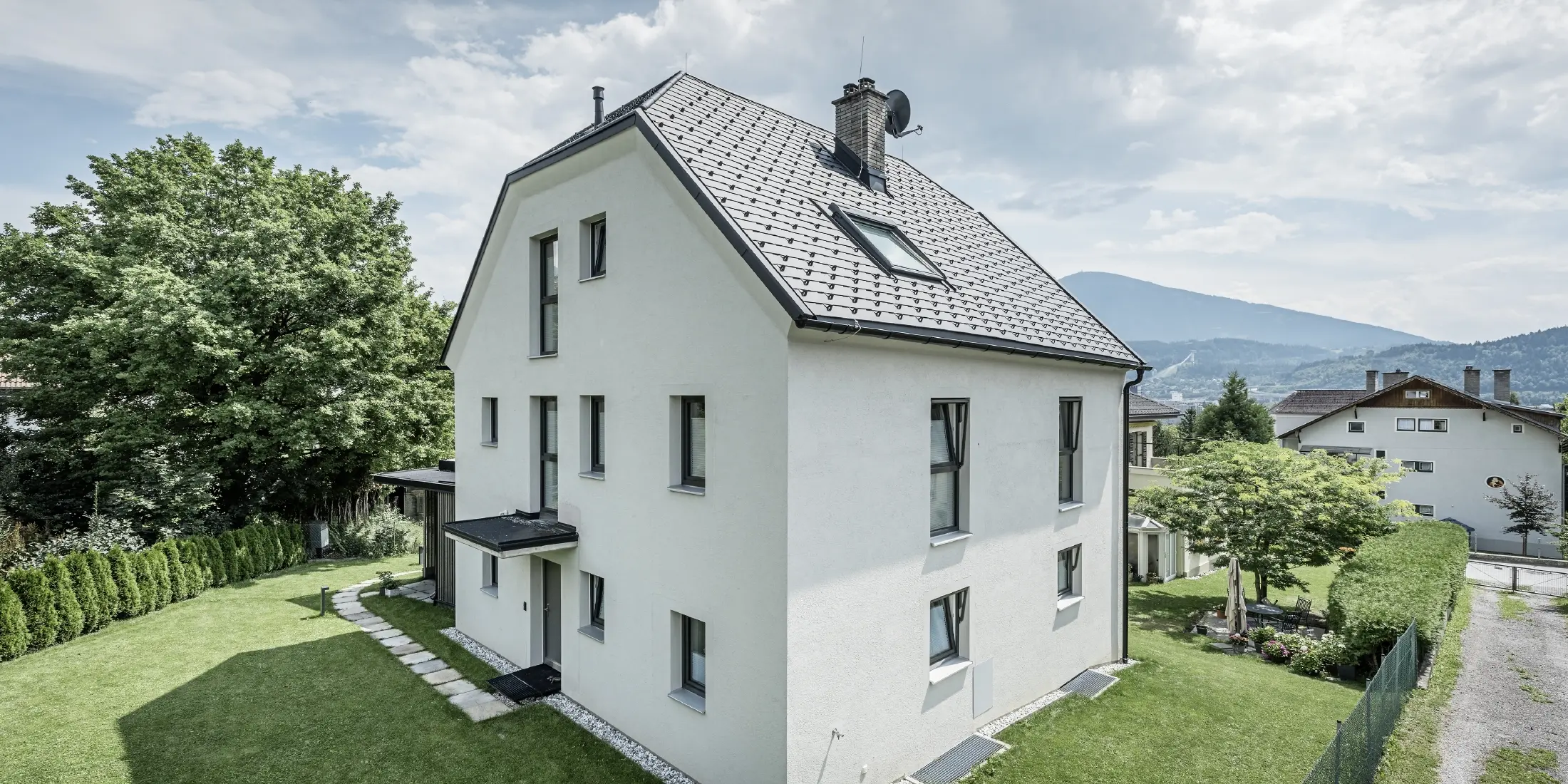 Modern three-storey detached house with a light-coloured plaster façade and dark grey aluminium gable roof from PREFA. The building has several narrow windows and anthracite-coloured roof drainage. It stands in a well-kept garden with a lawn, trees and hedges, with other residential buildings and a mountainous landscape visible in the background.