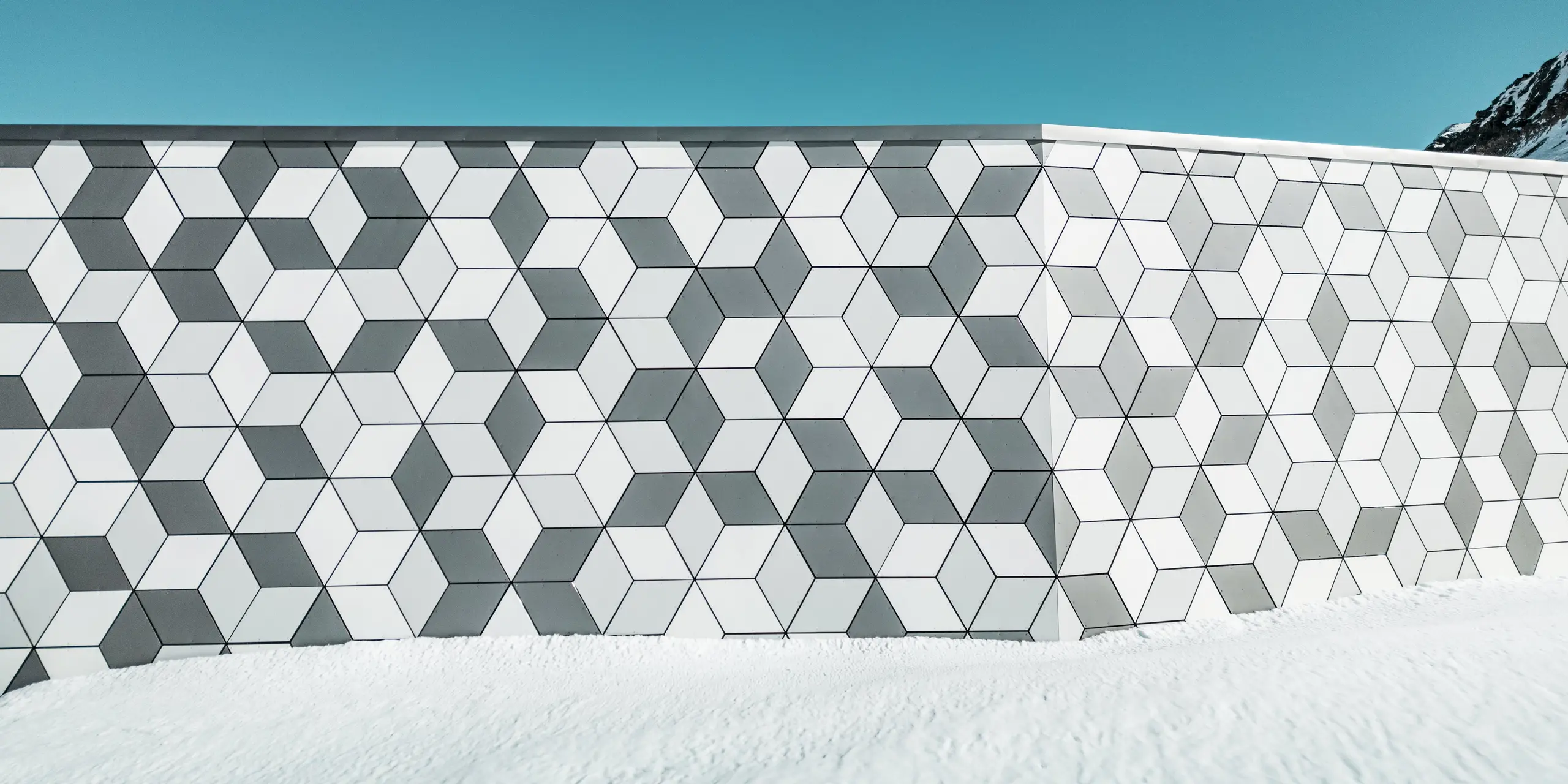 Die Nahaufnahme der Fassade der Seilbahnstation Jandri in Les Deux Alpes zeigt die präzise Anordnung der Rauten aus PREFABOND Aluminium Verbundplatten. Die angefertigten Rauten sind in einem auffälligen Muster angeordnet. Durch die harmonische Farbgestaltung in unterschiedlichen Weiß- und Grautönen fügt sich die Fassade perfekt in die schneebedeckte Alpenlandschaft ein. Die robusten und wetterbeständigen Platten sind eine langlebige und ästhetisch ansprechende Lösung für die extremen Witterungsbedingungen in den Bergen.