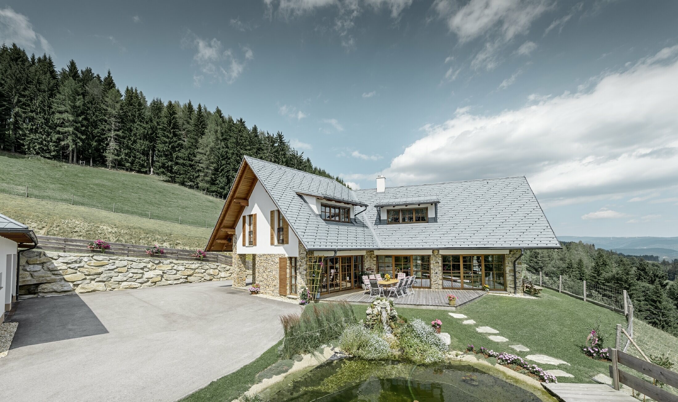 The detached house located on a slope, designed in an L shape, has a gabled roof with two eyebrow dormers. The roof was clad in PREFA roof shingles in stone grey. A lake is shown in the foreground and a forest can been seen in the background.