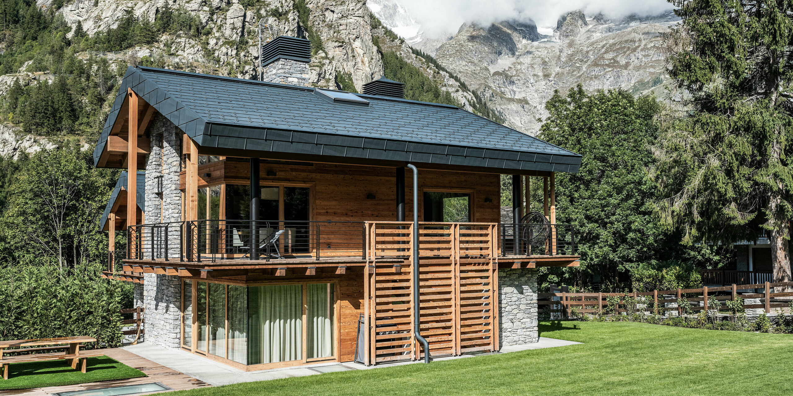 Attraktive Villa in Courmayeur mit einer Kombination aus Holz- und Steinmaterialien. An der Fassade und am Dach ist hochwertiges PREFA Blech zu finden. Die steingrauen FX.12 Aluminiumplatten fügen sich harmonisch in das beeindruckende Bergpanorama ein. Die Villa verfügt über einen großzügigen Balkon und eine Terrasse, die einen spektakulären Blick auf die majestätische Bergwelt bieten. Der Garten ist liebevoll angelegt und lädt mit seiner Rasenfläche zum Entspannen in der beeindruckenden Berglandschaft ein.