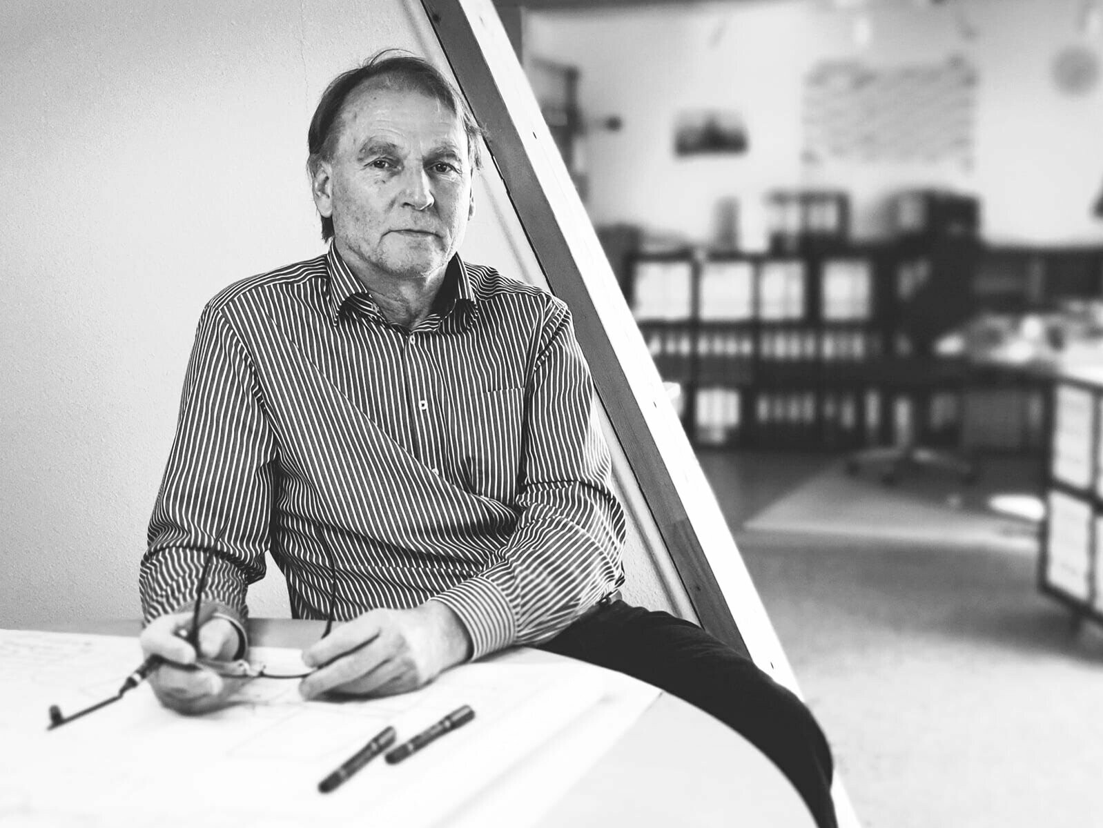 architect Andreas Geipel, black and white photo, is sitting at a round table and holding his glasses with a pen in his hand