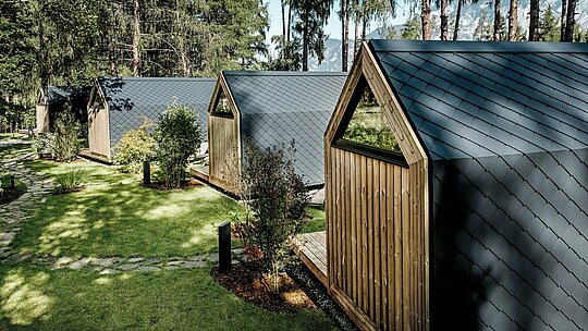 Eine Reihe minimalistischer Tiny Houses des Secret Koi Garden erstreckt sich eingebettet in eine gepflegte Grünanlage am Natterer See. Die geneigten Dächer und Fassaden sind mit kleinteiligen PREFA Aluminiumrauten in P.10 Anthrazit ausgeführt und erzeugen eine ruhige, fischhautartige Struktur. Umgeben von Bäumen und Sträuchern bieten die langlebigen, witterungsbeständigen PREFA Aluminiumoberflächen optimalen Schutz und verleihen dem Glamping‑Resort eine klare, zeitlose Architektursprache im Einklang mit der Natur.
