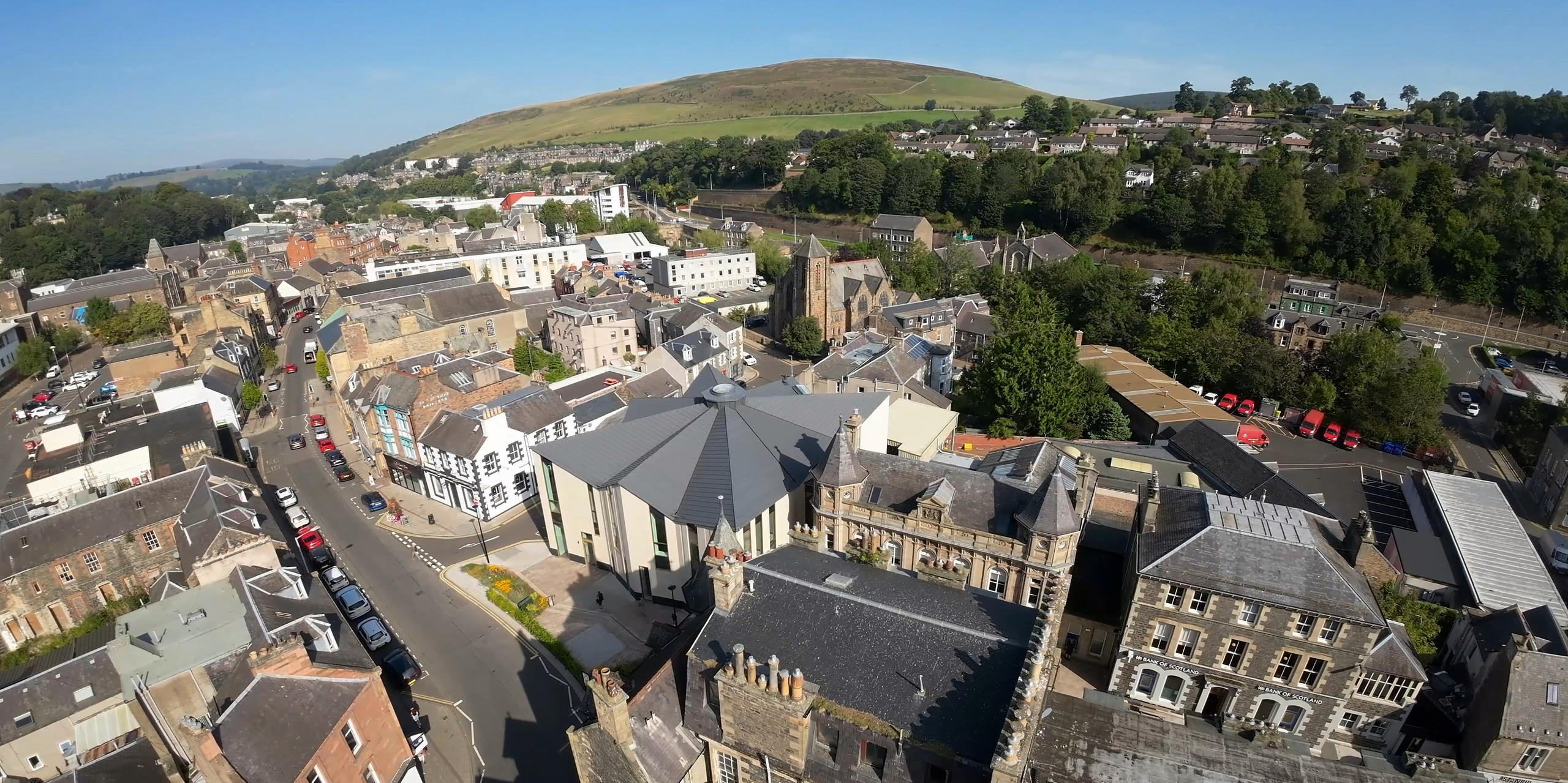Panoramic drone shot of 'The Great Tapestry of Scotland' gallery/visitor centre in Galashiels, framed by the picturesque Scottish hills. The striking building stands out with its PREFALZ roof system in P.10 Zinc Grey, which curves elegantly over the unique structure. The unusual lines of the roof clearly stand out from the surrounding traditional buildings and show how PREFA aluminium products combine modern architecture with the city's cultural heritage.