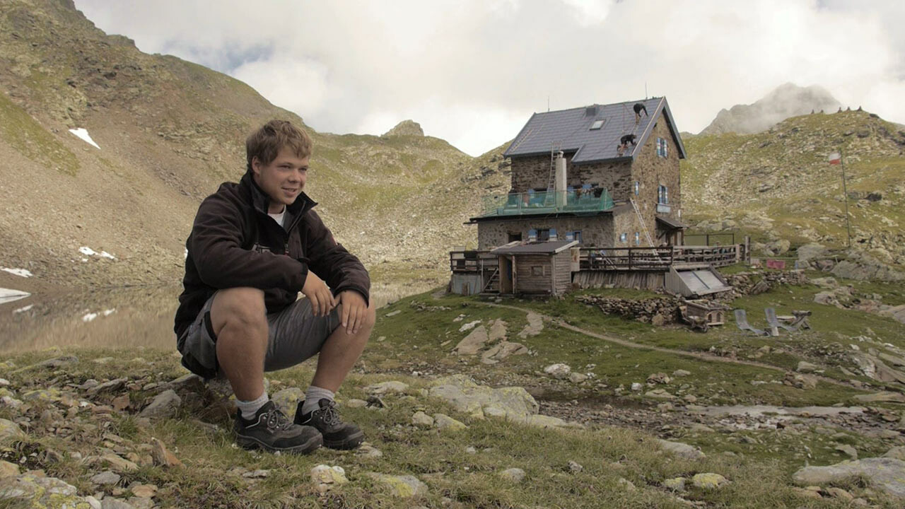 The apprentice Jakob Schöttl sits on a stone. In the background you can see the "Flagger Schartenhütte", which has been re-roofed with PREFA roof panels.