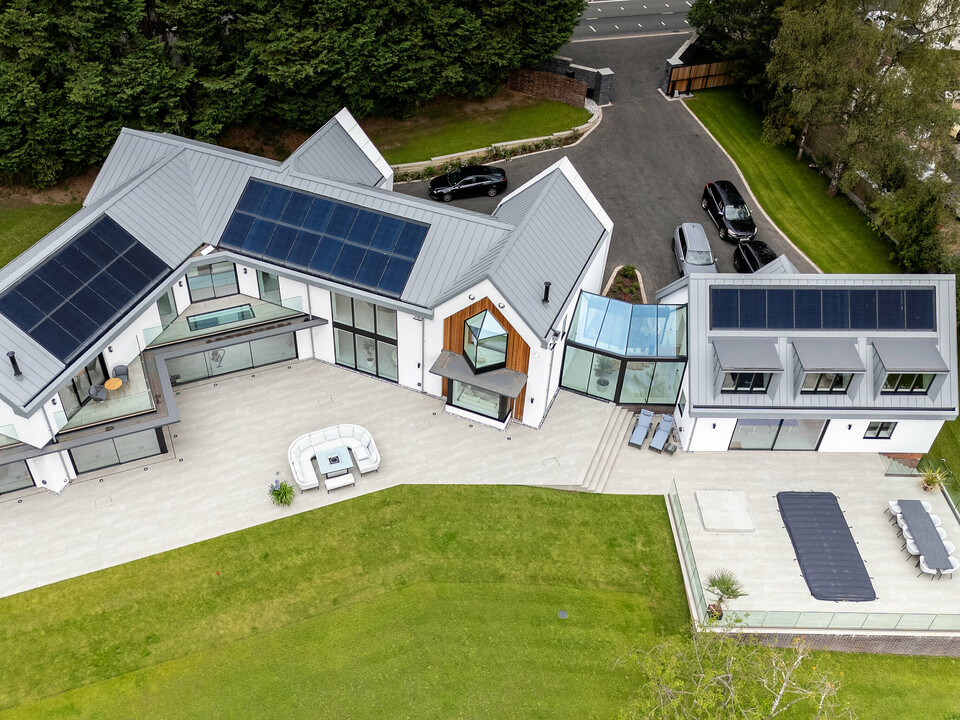 Das Bild zeigt eine beeindruckende Vogelperspektive der modernen Villa in Draycott in the Clay mit ihrem markanten PREFALZ-Dach in der Farbe Patinagrau. Die Stehfalzdeckung des Daches setzt sich harmonisch von der umliegenden grünen Landschaft ab und unterstreicht das moderne, elegante Design des Hauses. Die nahtlos in das Dach integrierten Solarpanels betonen die energieeffiziente Ausrichtung des Hauses. Die großzügige Terrasse und der gepflegte Garten vervollständigen das Bild dieser innovativen Architektur. Durch die Verwendung von PREFALZ ist höchste Qualität und Langlebigkeit garantiert.