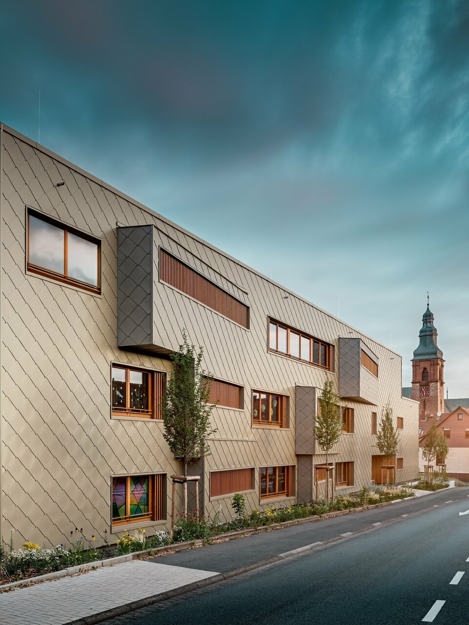 Im sanften Licht des Sonnenuntergangs zeigt sich die Kindertagesstätte in Alzenau von ihrer stimmungsvollsten Seite. Die Fassade aus bronzefarbenen PREFA Wandrauten 44 × 44 reflektiert das Abendlicht und betont die hochwertige, strukturierte Oberfläche des modernen Hybridgebäudes. Die klar gegliederte Architektur mit versetzten Fensterbändern und vertikalen Holzlamellen steht in spannendem Kontrast zur historischen Kirche im Hintergrund. Links im Bild sorgt ein filigranes Metallgerüst für leichte Transparenz beim Außentreppenhaus. Der gepflegte Grünstreifen entlang des Gebäudes unterstreicht den ökologischen Anspruch. Dieses Bild verbindet eindrucksvoll moderne Baukultur, nachhaltige PREFA Aluminiumtechnologie und harmonische Einbettung in das historische Stadtbild von Alzenau.