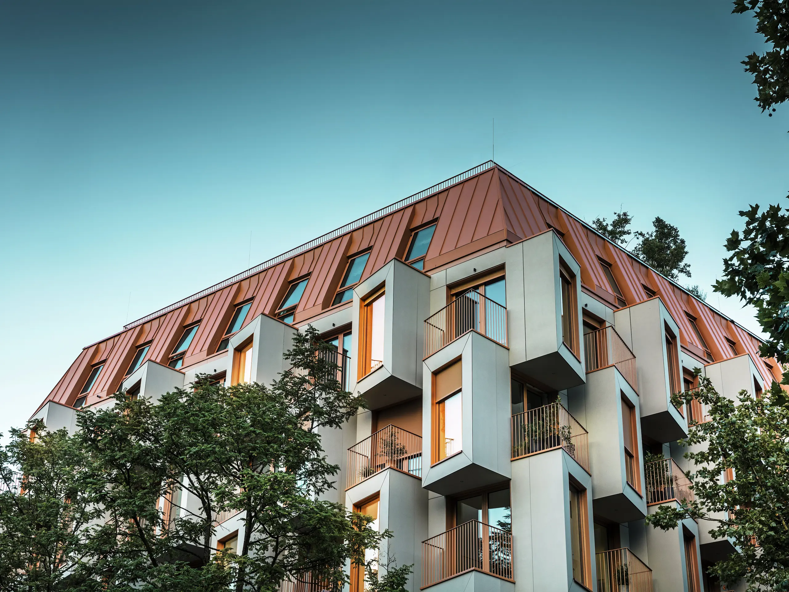 Architecturally outstanding façade of the Van B residential project in Munich with striking bay windows and PREFALZ aluminium roof in the special colour IGP Orange HDP. At dusk, the view from below reveals the lights in the windows. The modern roof solution combines maximum weather resistance with an elegant appearance and adds colourful accents to the urban landscape. The interplay of glass fibre reinforced concrete, copper-orange window frames and aluminium roof creates a dynamic, durable and sustainable building envelope – ideal for future-oriented construction.