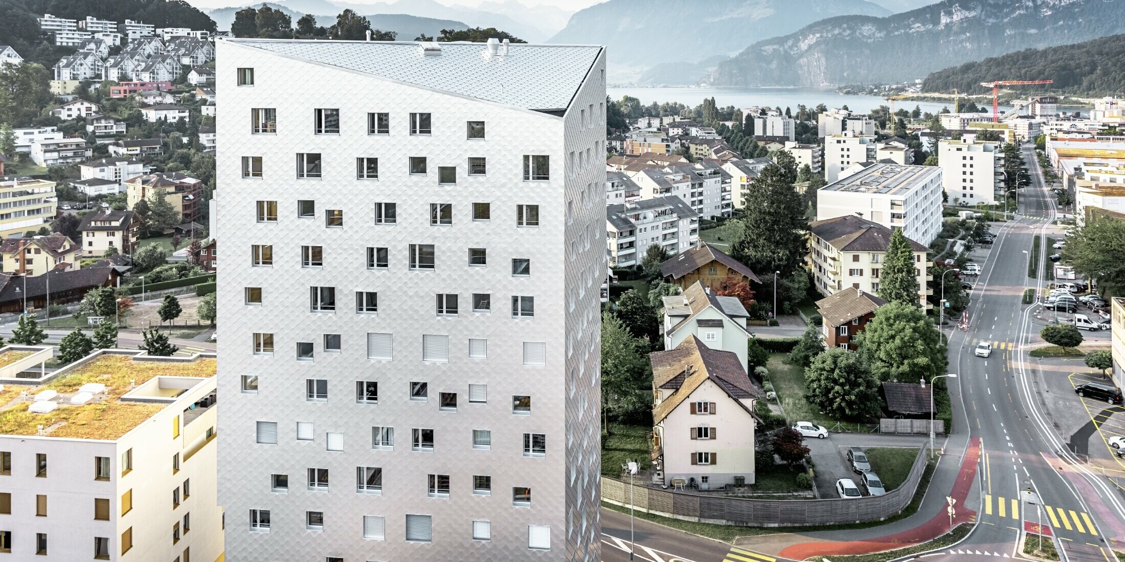 Das Bild zeigt das Solitaire-Hochhaus in Horw, einem Vorort von Luzern, das von der Schweizer Architektin Tilla Theus entworfen wurde. Das Hochhaus hat eine Fassade aus eloxierten Aluminiumrauten, die die Umgebung und die Wolken reflektieren. Die Fenster sind unregelmäßig angeordnet und als 'tanzende Fenster' bekannt. Im Hintergrund sind weitere Gebäude, Straßen und die malerische Berglandschaft der Schweiz zu sehen. Das Hochhaus steht inmitten einer städtischen Umgebung mit einer Mischung aus Wohnhäusern und städtischer Infrastruktur.