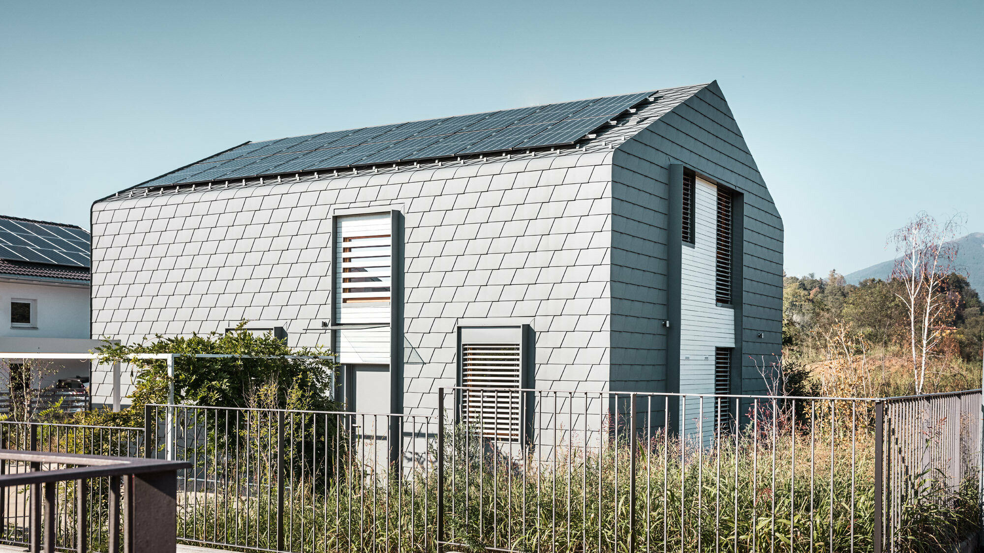 Lateral view of "Casa Longaretti" with a green area and fence, the round transition between roof and façade cis clearly visible, just like the light grey shingle façade.