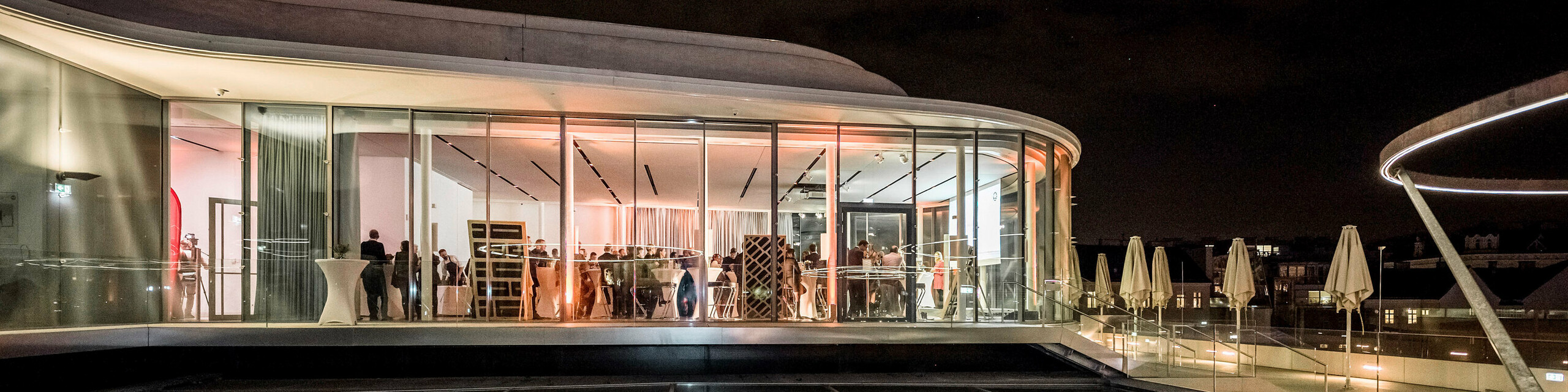 Night shot of the Libelle MQ on the roof of the Leopold Museum with a reflecting surface, the guests are inside the illuminated event space.