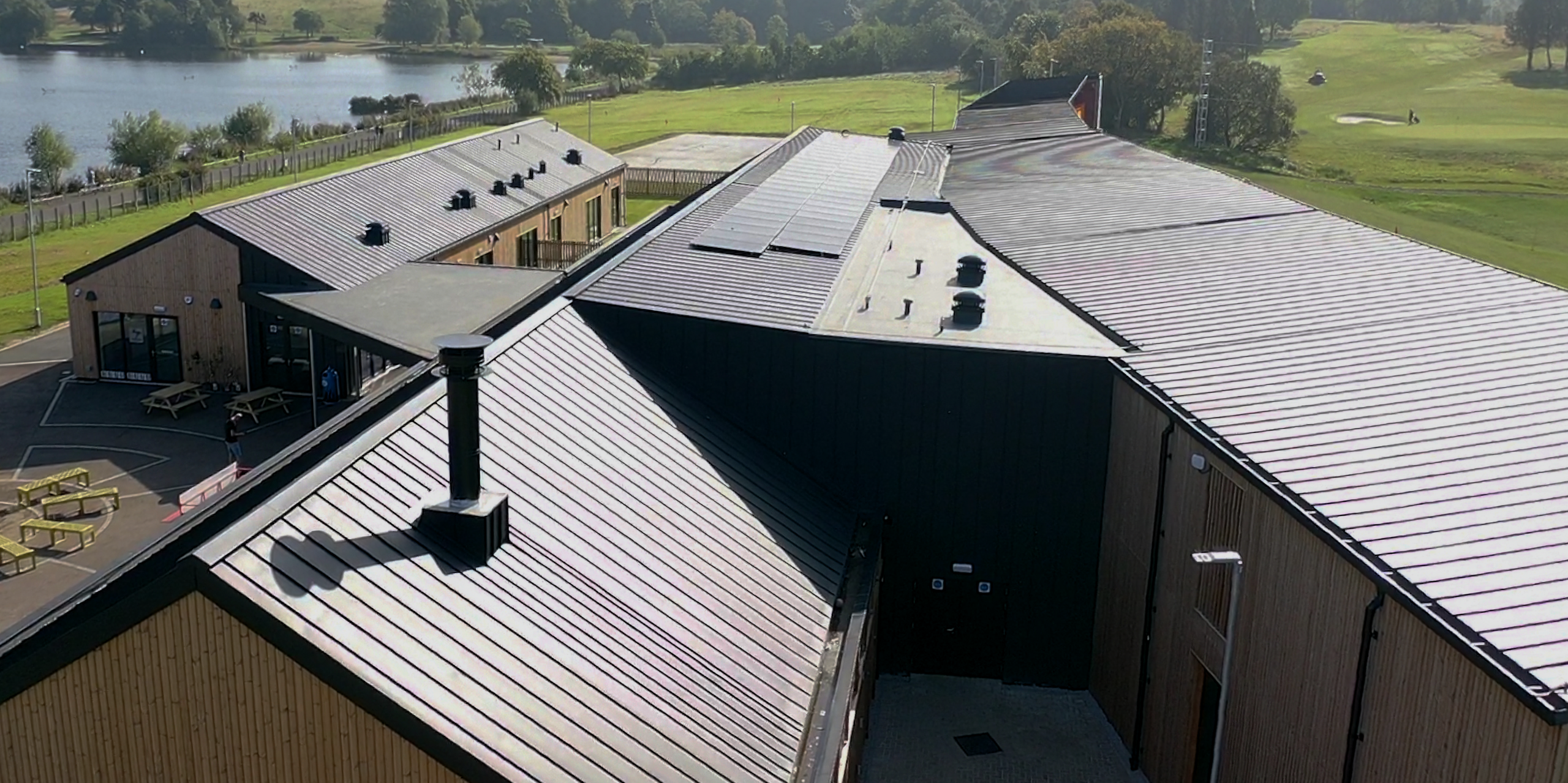 Aerial view of the Golf it! Driving range – golf venue in Glasgow, Scotland, shows the precisely installed PREFALZ roof system in P.10 black. The extensive facility, with a roof area of around 2,000 square metres, reflects the high quality of PREFA aluminium products. The roof surface blends harmoniously into the idyllic, green landscape with the neighbouring lake, emphasising the importance of functionality and natural beauty. The PREFA gutters round off the elegant appearance and guarantee durability and reliability in all weathers.