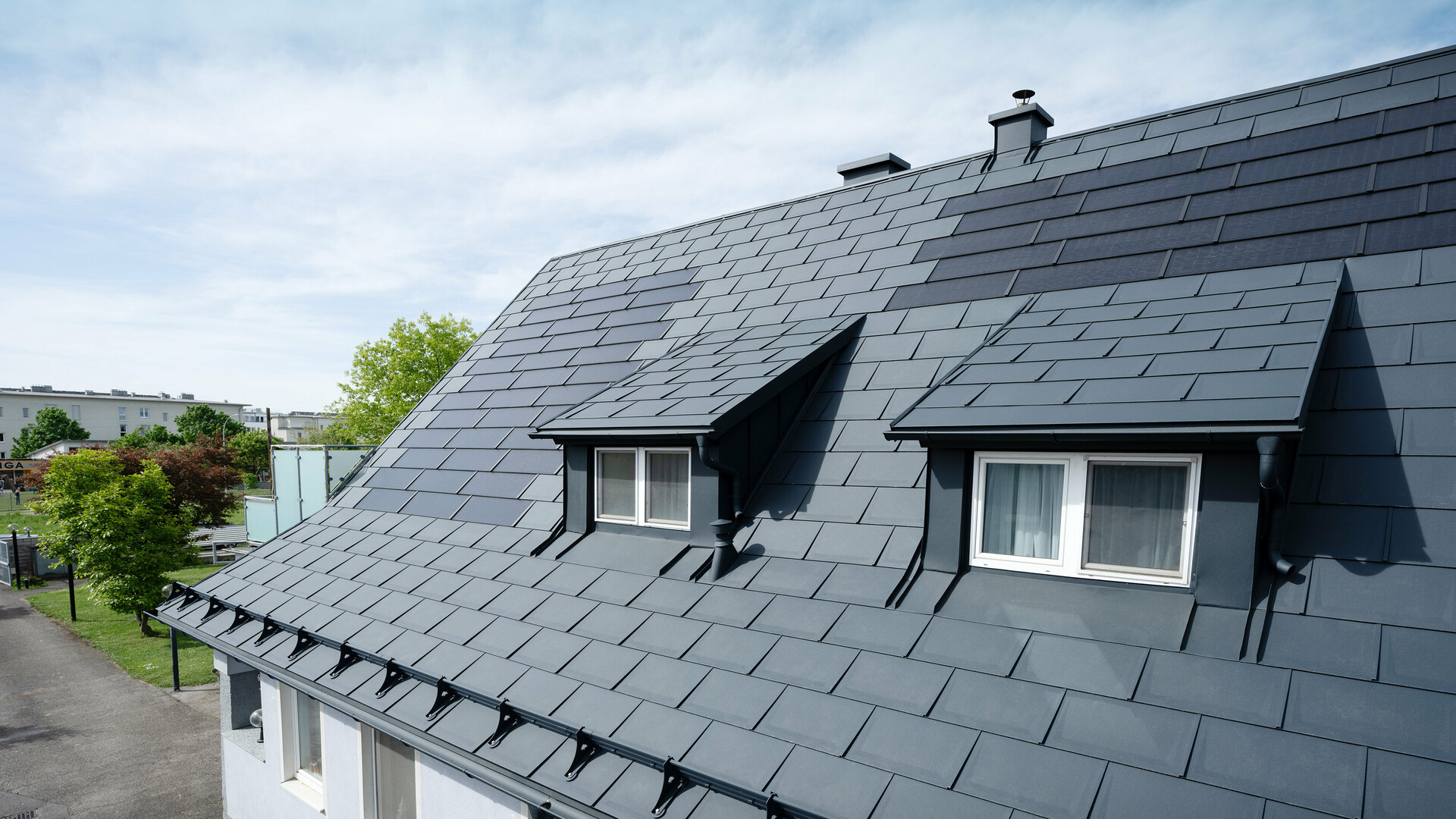 A close-up of a roof covered with large PREFA solar roof panels and the PREFA R.16 roof tile in P.10 anthracite. Two dormers are visible. The solar roof panels are evenly distributed across the roof, providing a modern, sustainable solution.