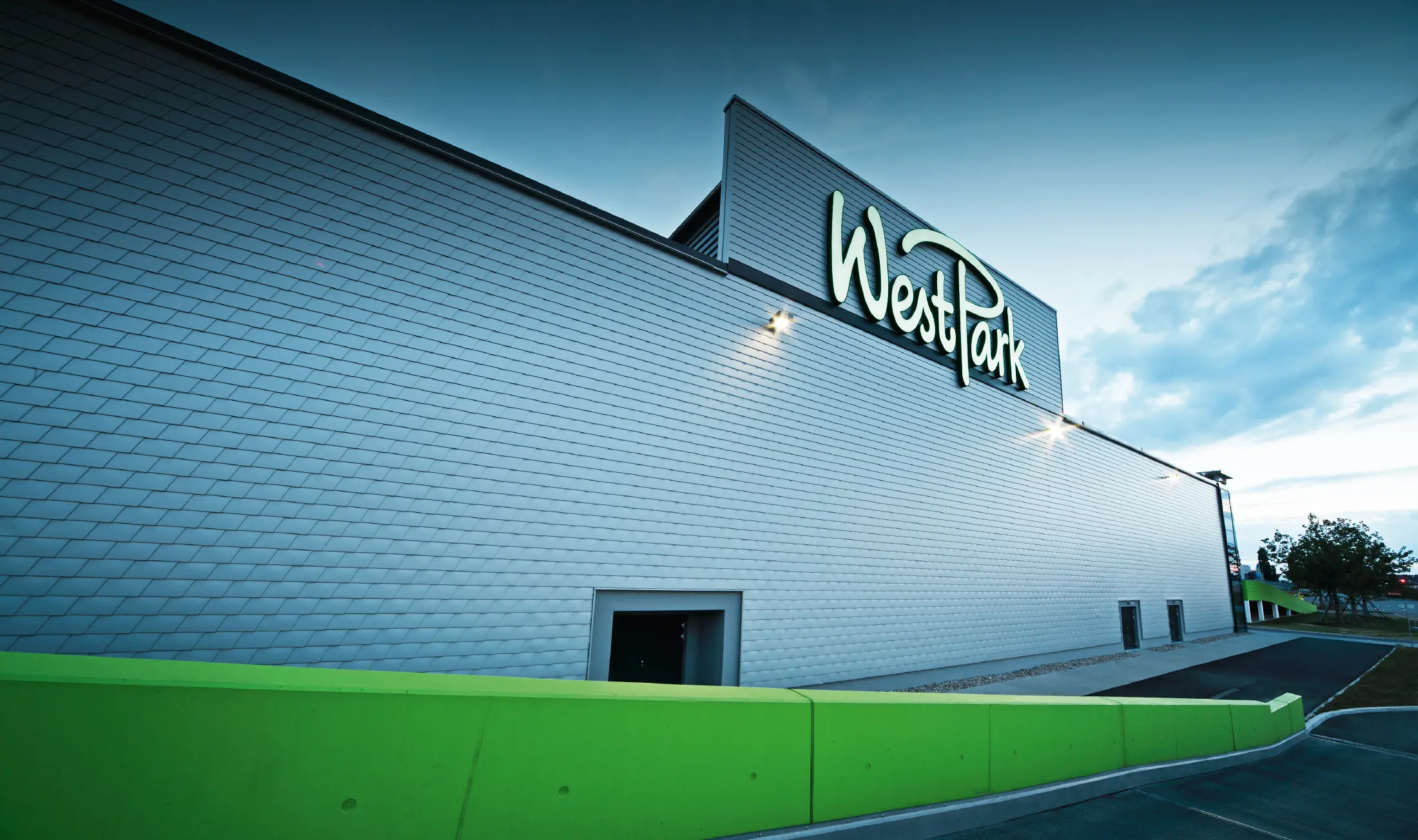 Moderne Fassade eines Einkaufszentrums mit großem Schriftzug „WestPark“. Die Außenwand ist mit silbermetallicfarbenen Aluminium-Schindeln verkleidet und wirkt durch die gleichmäßige Struktur elegant und technisch. Im Vordergrund verläuft eine leuchtend grüne Mauer entlang des Gehwegs. Das Gebäude ist in der Abenddämmerung beleuchtet.