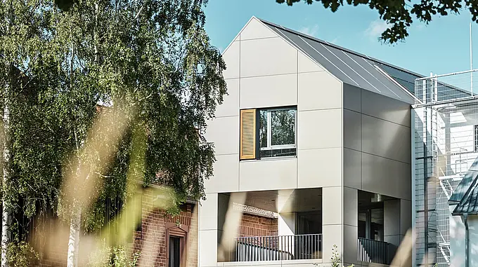 The image shows the modern town hall in Alzenau, whose façade is made of aluminium composite panels from PREFA in an elegant bronze tone. The pointed roof area and large windows emphasise the contemporary character of the building. In the foreground, trees and green spaces frame the building, while the soft lighting and clear sky underline the modern urban space.