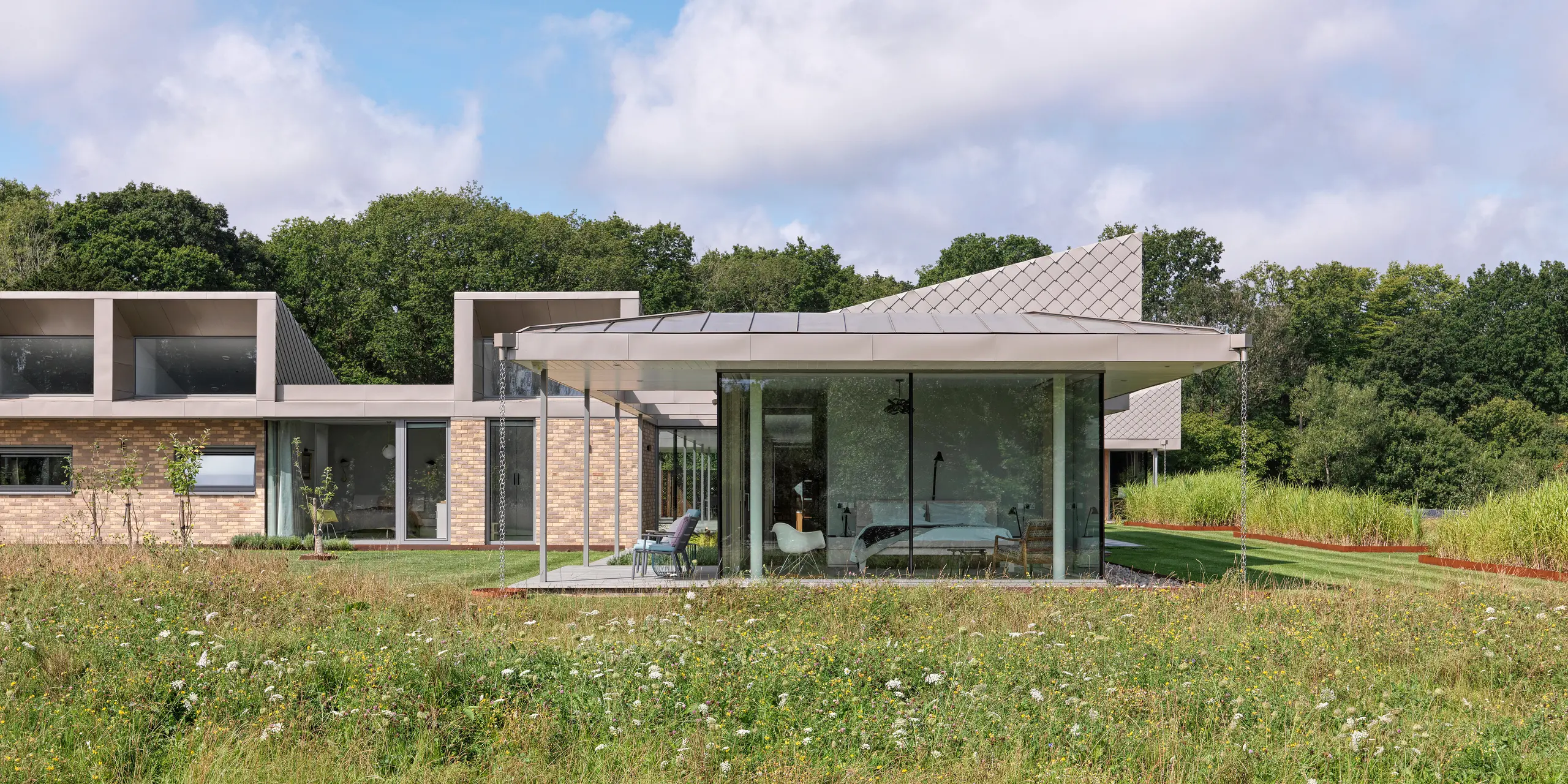 Side view of the modern bungalow in West Sussex with standing seam roofing and accentuated façade areas made of PREFA rhomboid façade tiles 20 × 20 in P.10 bronze.The floor-to-ceiling glass fronts of the living areas open onto a wildflower meadow in the foreground, creating a seamless connection between the interior and nature. The durable PREFA roof and façade system not only protects the building, but also lends it architectural elegance.